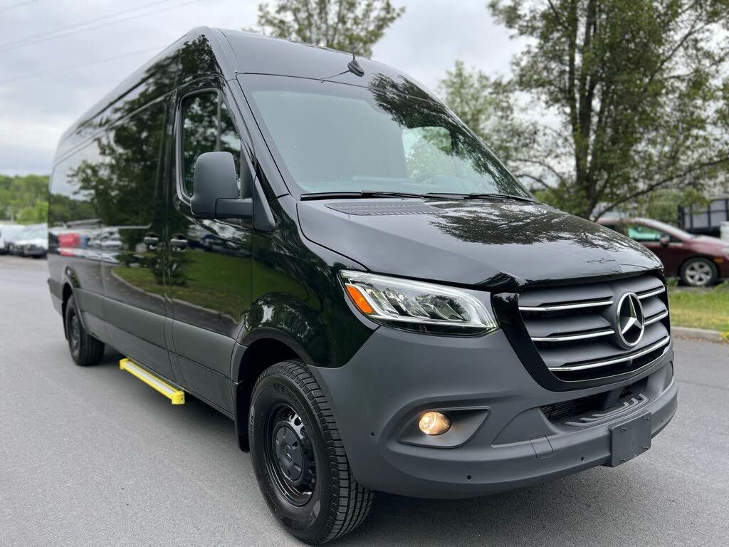 Black Mercedes-Benz Sprinter van parked on a paved road. Gray bumper, black tires, and yellow side step.