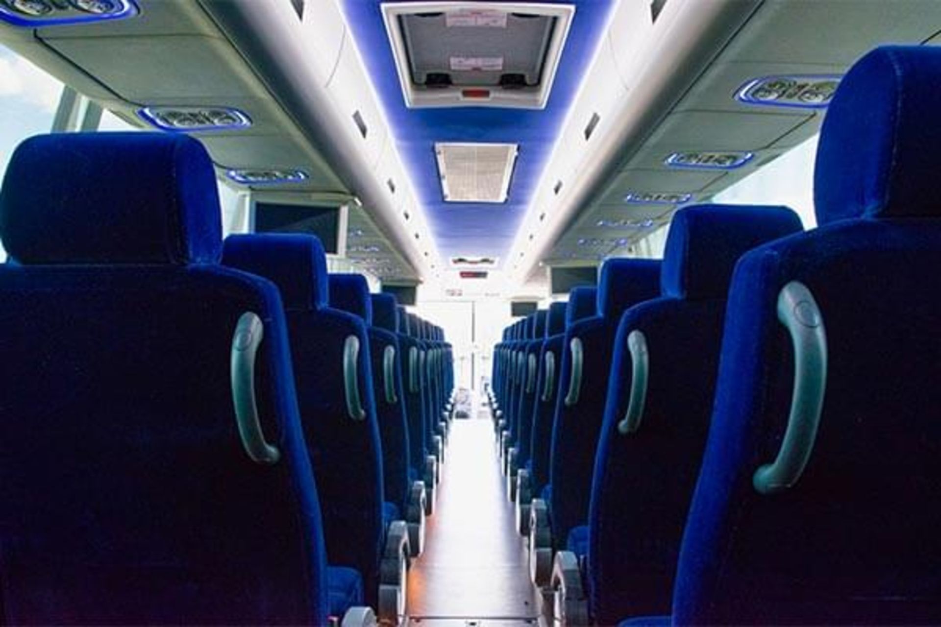 Interior view of a bus with rows of blue seats. The ceiling is lit with blue lights and there are windows in the distance.