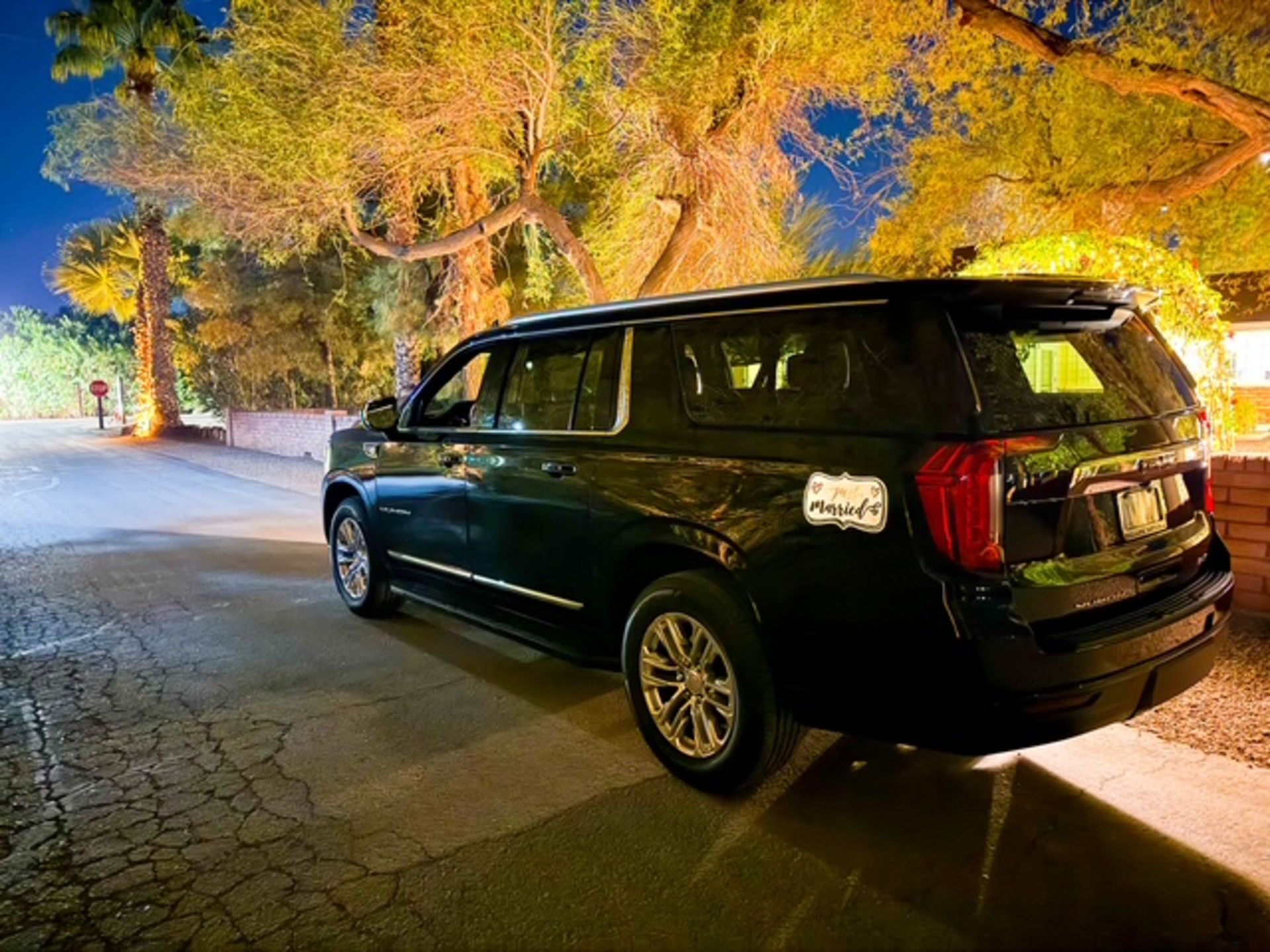 Black SUV parked on a cracked pavement at night, near a building, trees and street sign illuminated.
