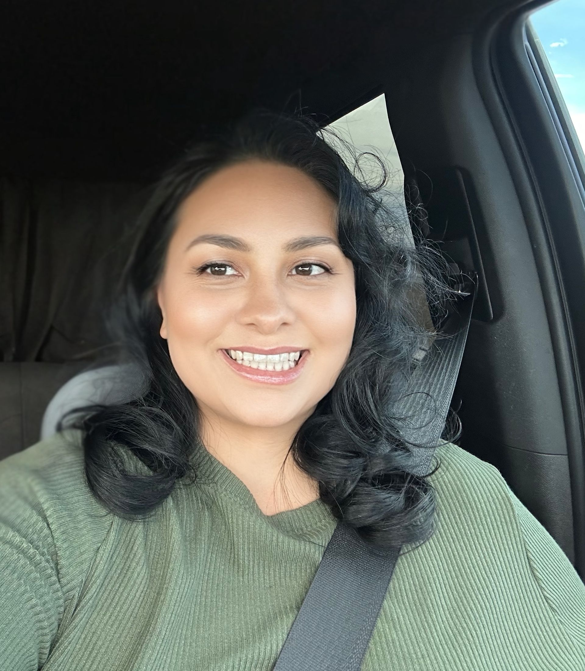 Woman with dark hair smiles broadly while seated in a car, wearing a green sweater, with a seatbelt on.