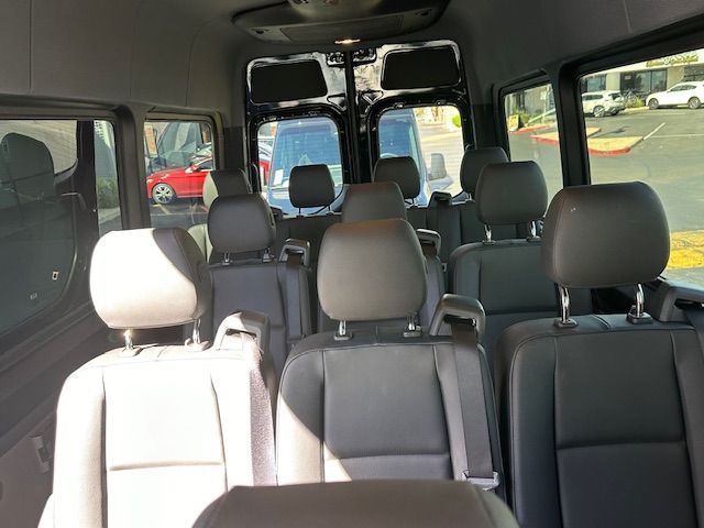Interior view of a van with several rows of black seats, some facing forward. Bright sunlight streams in from windows.