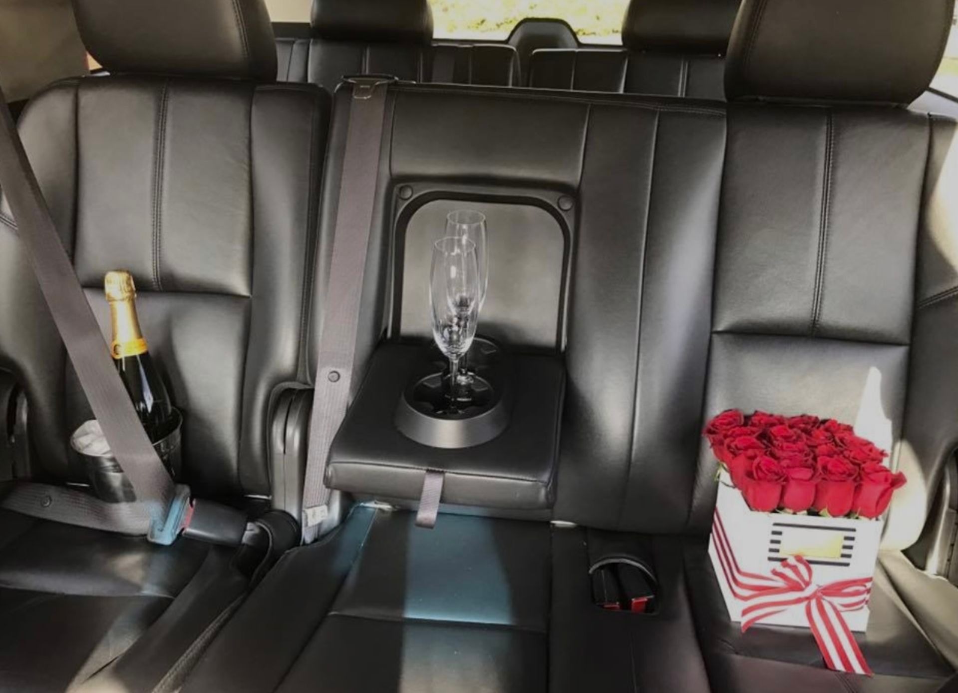 Backseat of a car with a champagne bottle, glass, and a box of red roses. Black leather seats; luxury setting.