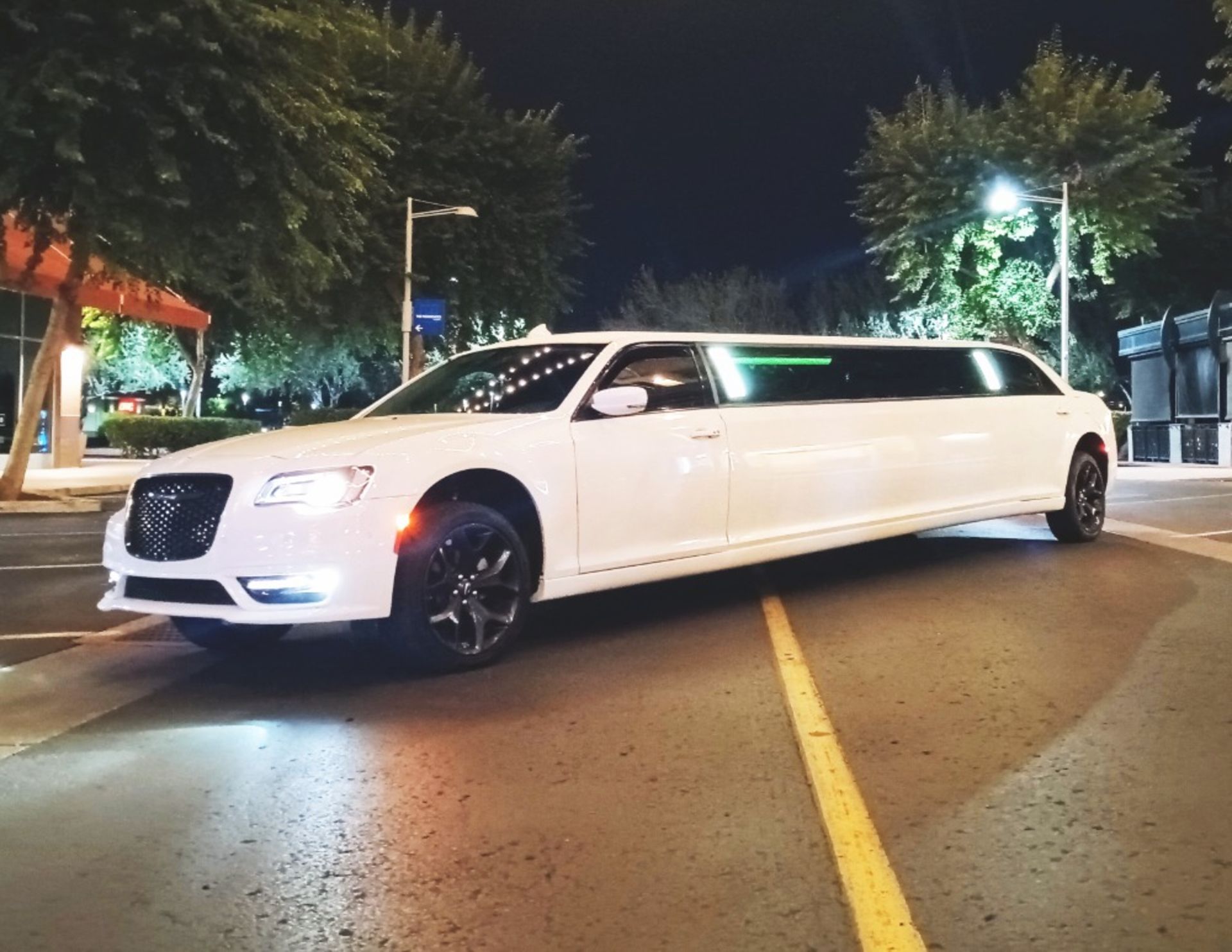 White limousine parked on a city street at night, headlights on. Trees and streetlights are visible in the background.