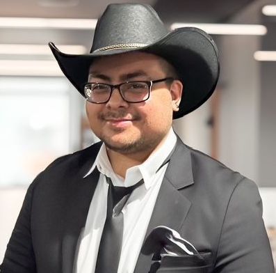 Man in a black cowboy hat, suit, glasses, and tie; smiles at the camera. Office background.