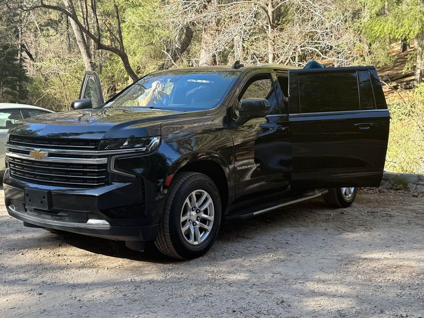Black Chevrolet SUV parked on gravel with driver's side door open. The setting is outdoors, trees visible in the background.