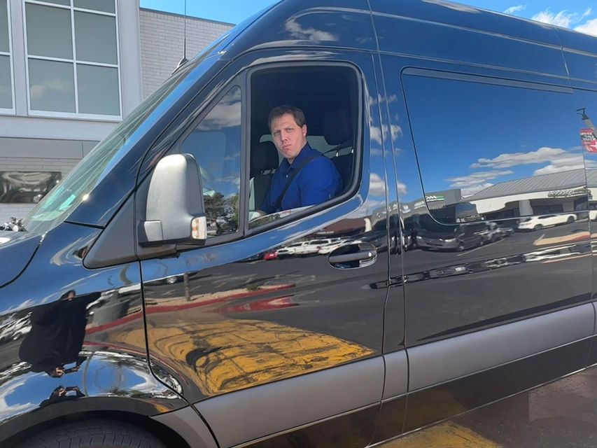 Man in blue blazer seated in the driver's seat of a black van, looking directly at the camera.
