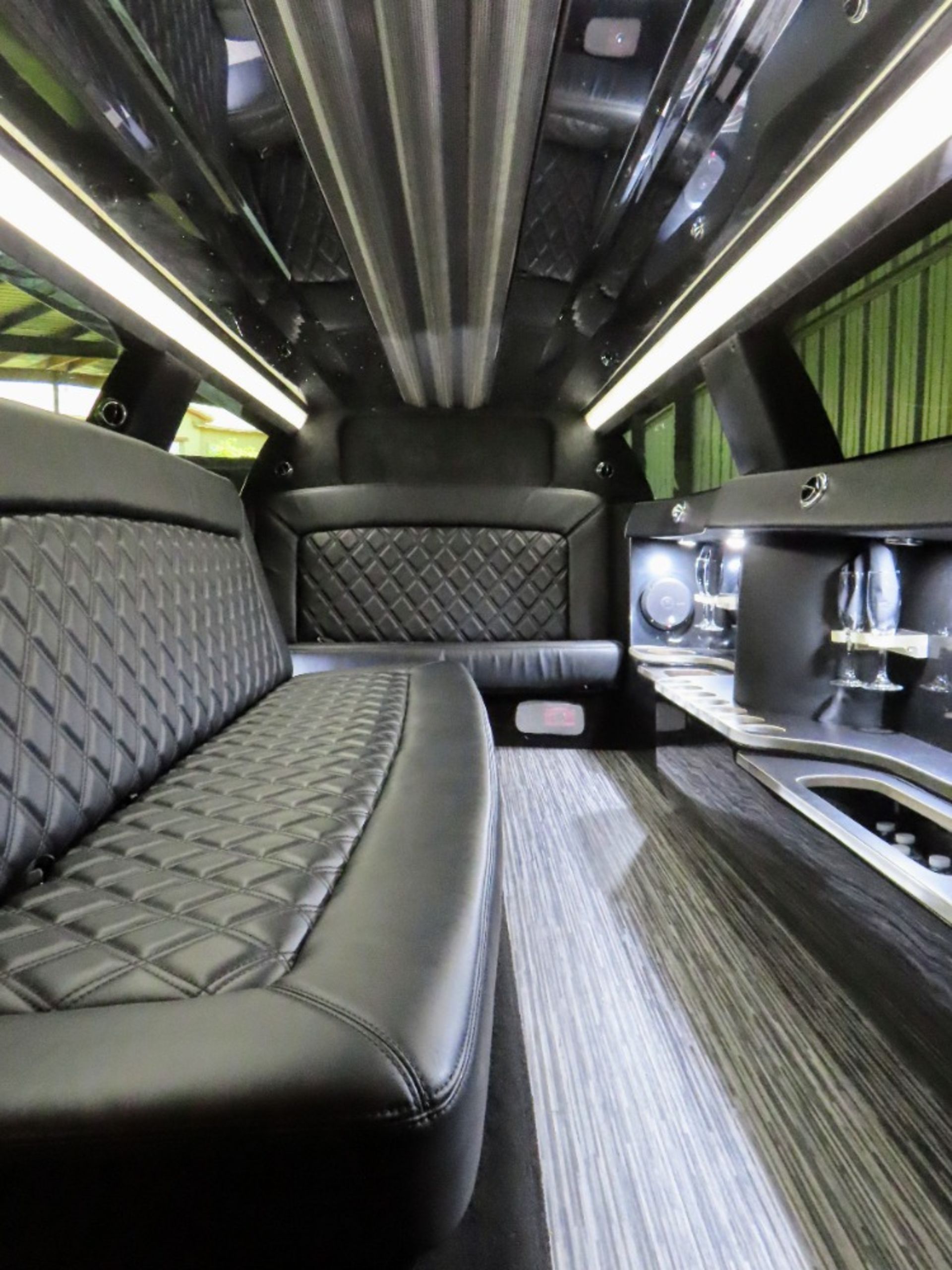 Interior of a black limousine with diamond-stitched leather seating, bar area, and wood-look flooring; overhead lights.