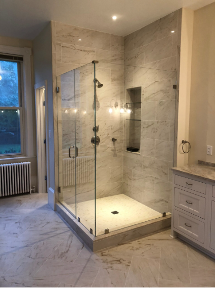 A glass-enclosed shower with light marble-patterned tile walls and floor, located in a bright bathroom.