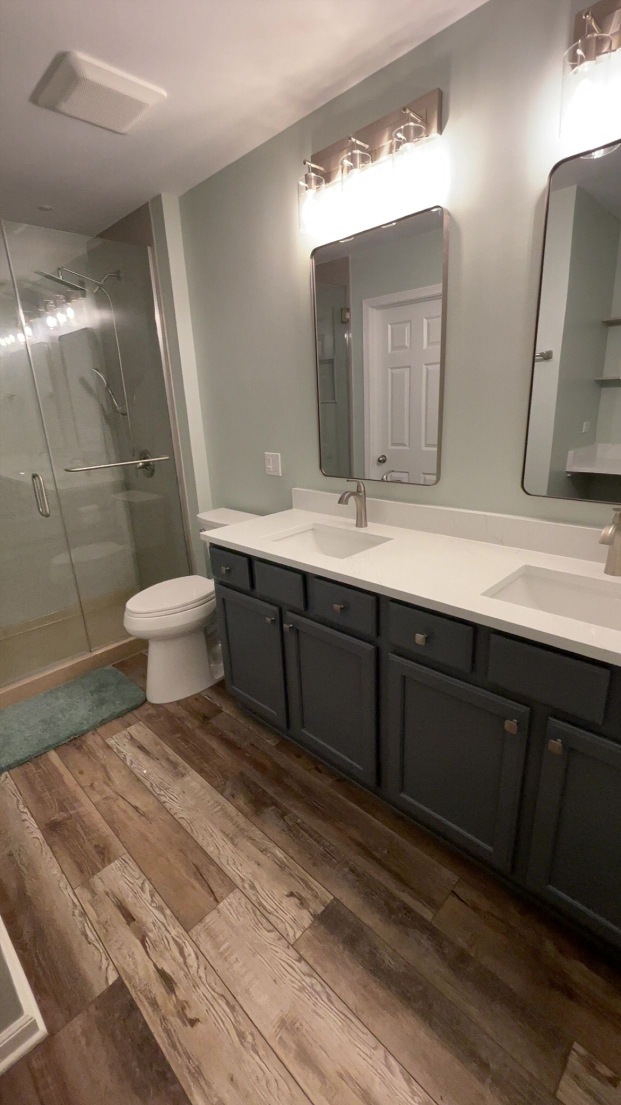 Bathroom with gray cabinets, white countertop, two mirrors, shower, and wood-look flooring.