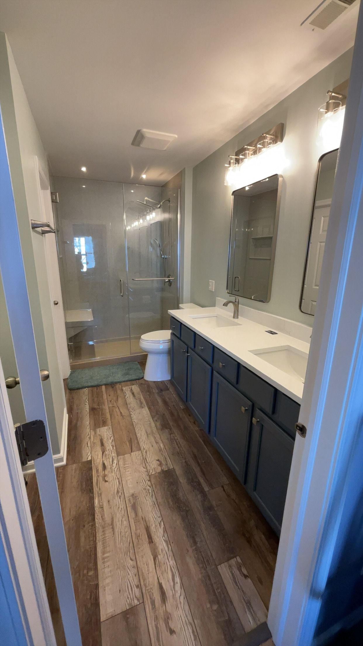 Bathroom with a blue vanity, wood-look flooring, glass shower, and two silver-framed mirrors.