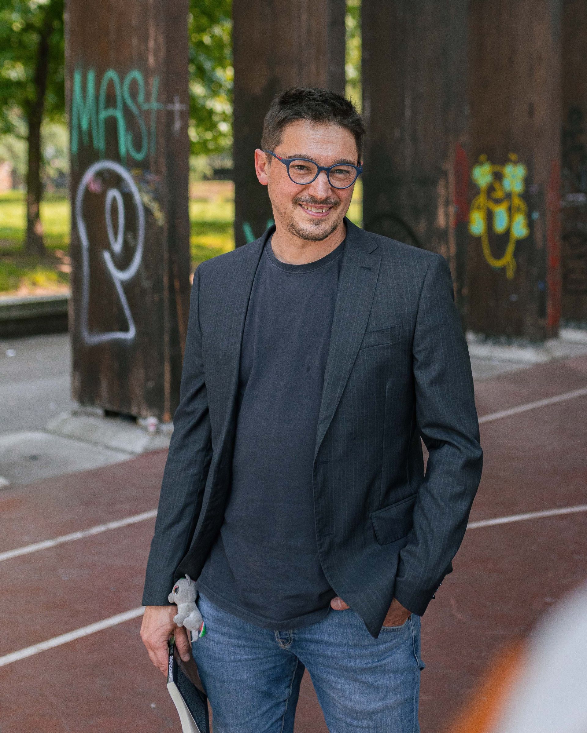 Un uomo con occhiali e blazer sorride, in piedi vicino a pilastri coperti di graffiti in un'area all'aperto.