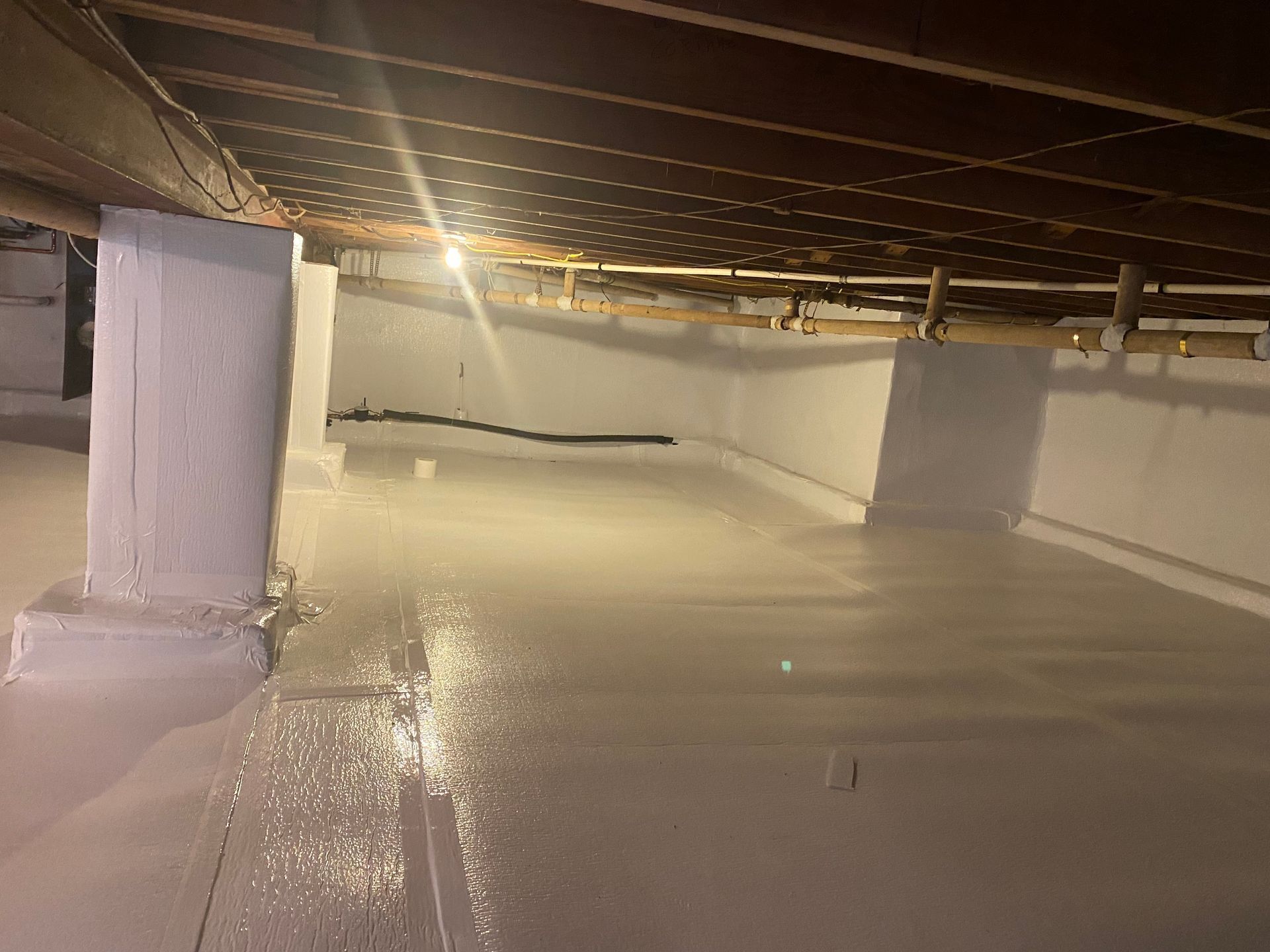 Person spraying a crawl space with white liner on walls and floor. Pipes and beams overhead.