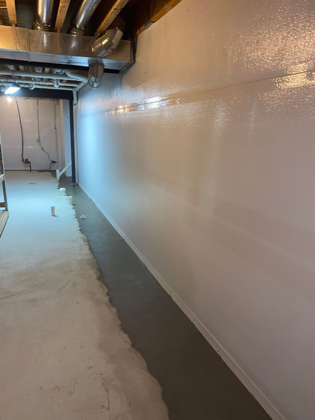 A sump pump system in a gray-painted basement, including a pump, piping, and a black backup power unit.