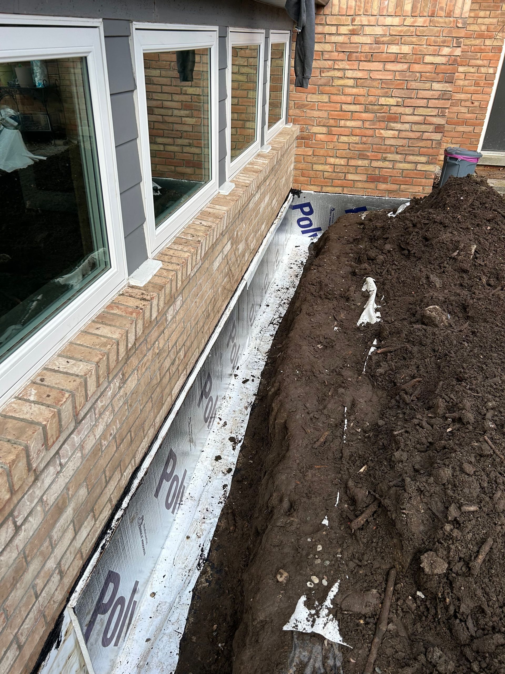 Workers waterproofing a foundation wall, applying black sealant.  Trench dug alongside the house; heavy equipment visible.