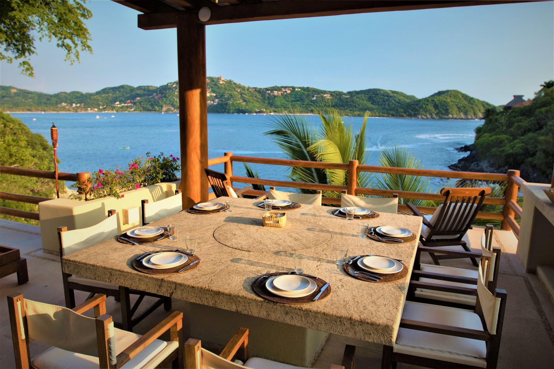 Mesa de comedor al aire libre para ocho personas con vista a la costa del agua y a las colinas verdes.