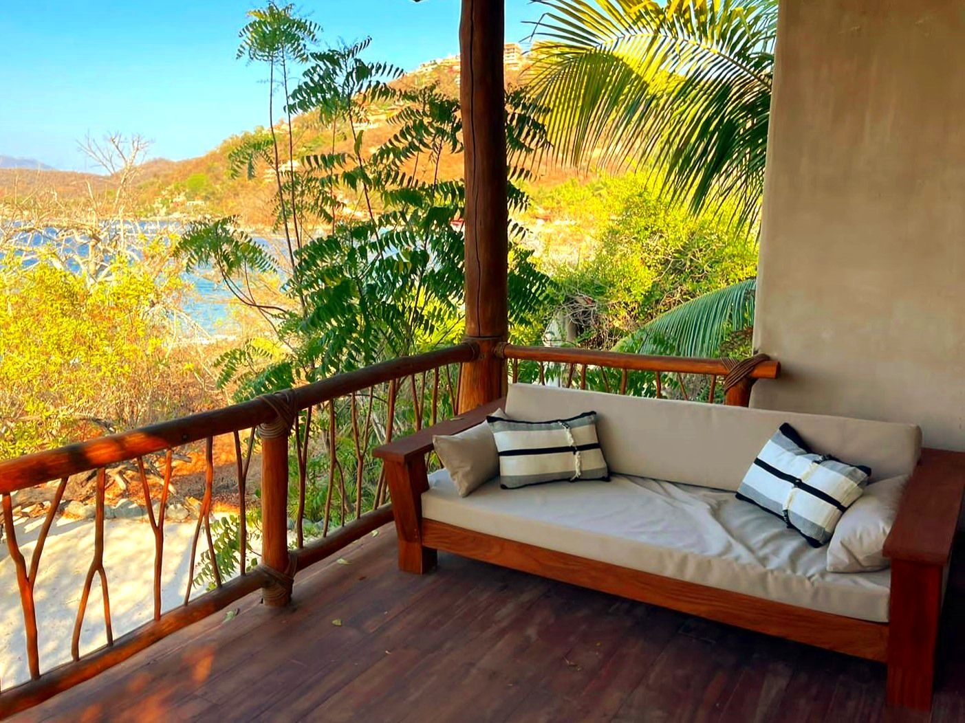 Porche de madera con sofá con vista a una bahía y exuberante vegetación bajo un cielo soleado.