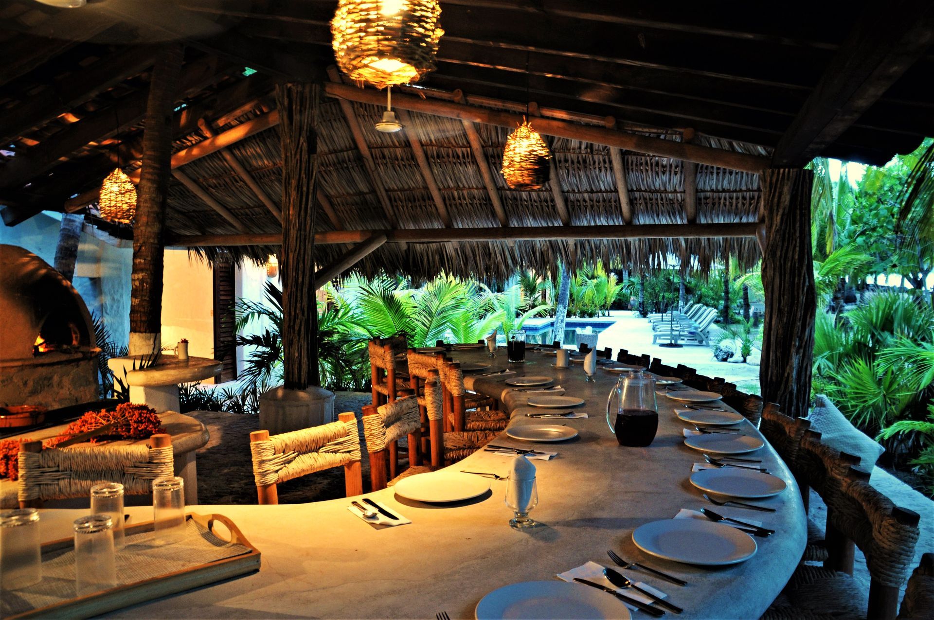 Comedor al aire libre con una mesa larga preparada para comer, bajo un techo de paja, rodeado de exuberante vegetación.