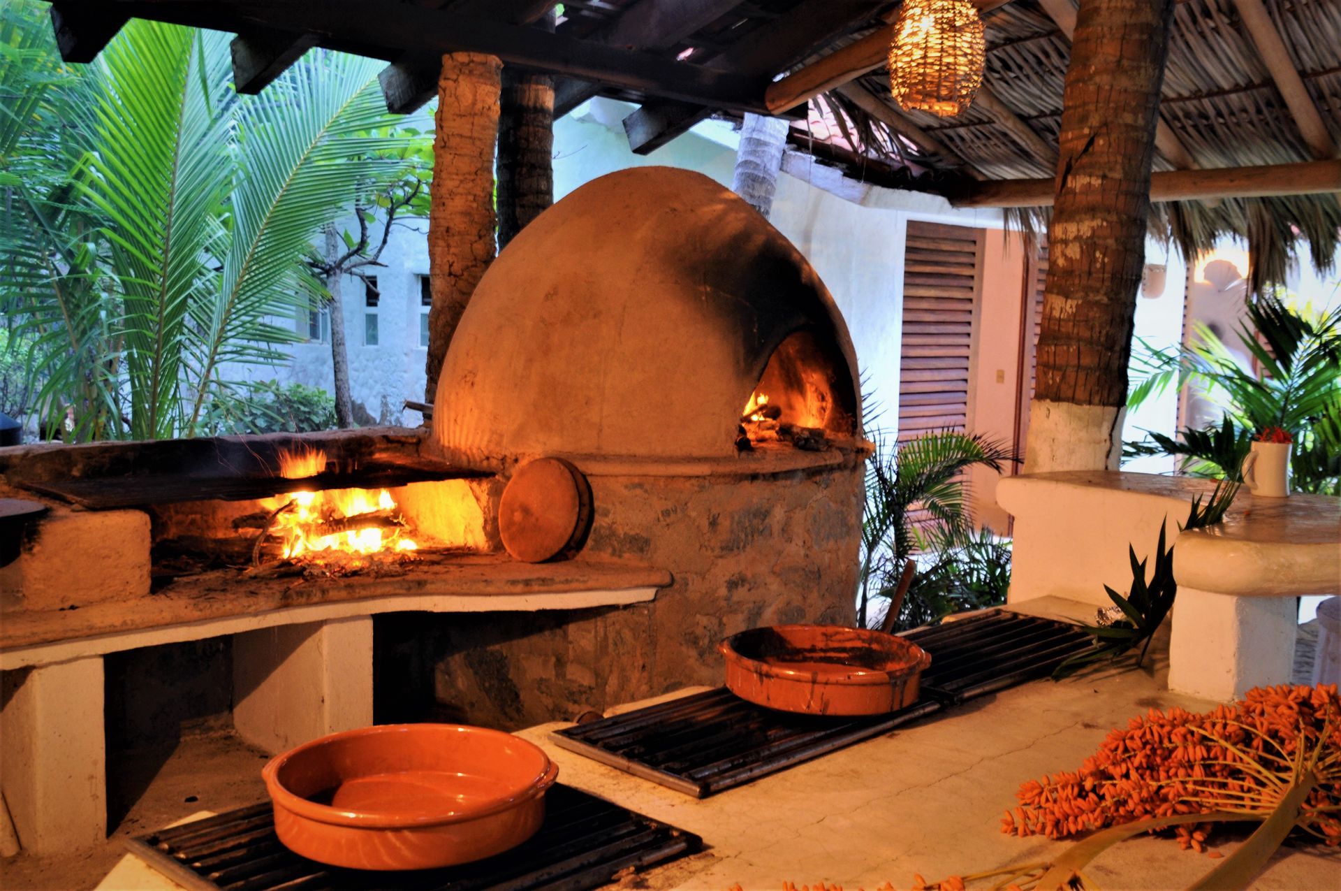 Cocina al aire libre con horno de leña, parrilla y vajilla de terracota; palmeras al fondo.