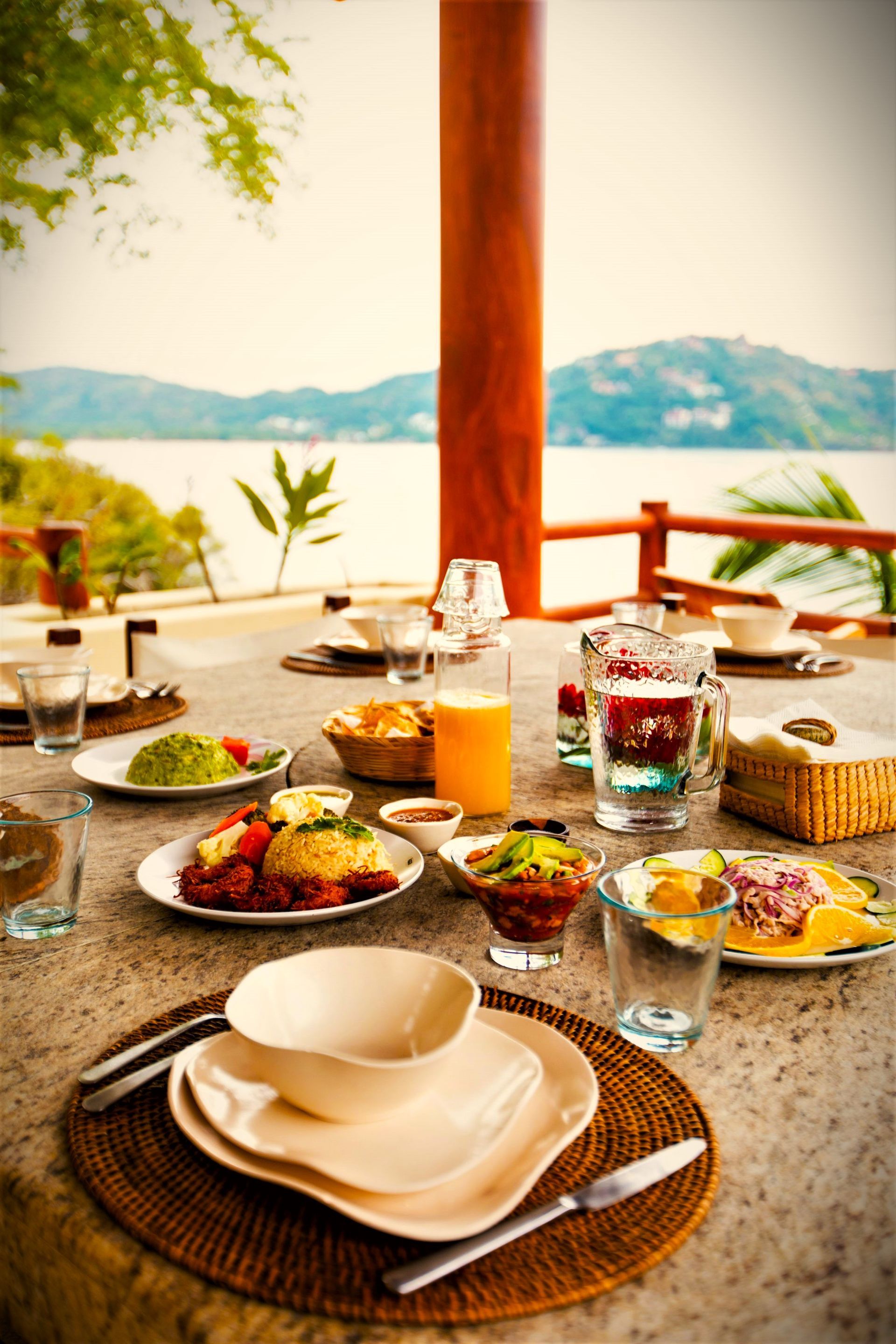 Mesa al aire libre preparada para comer, con vista al mar de fondo. Platos, bebidas y vasos.
