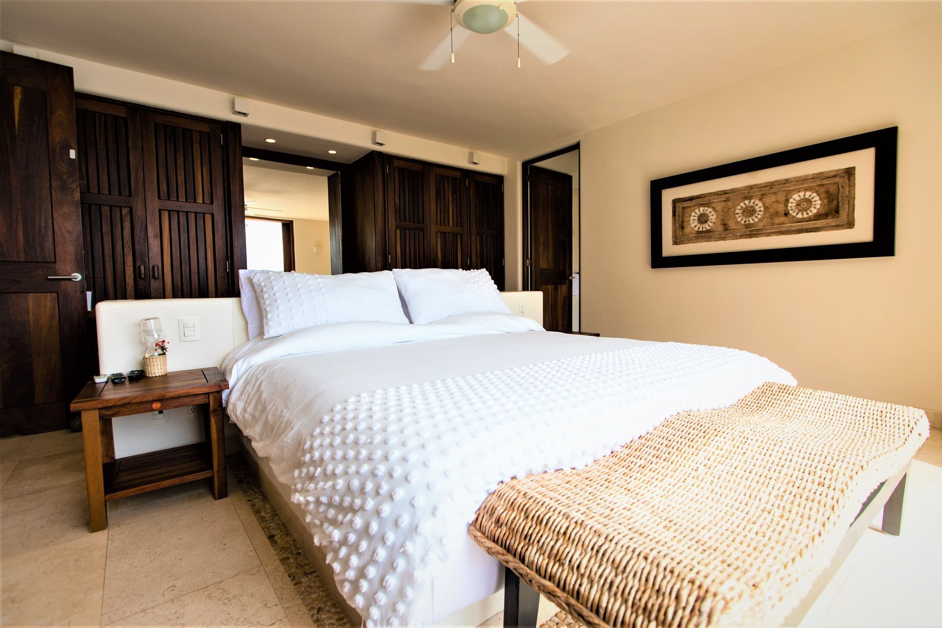 Dormitorio con cama blanca, detalles de madera y un banco de mimbre.