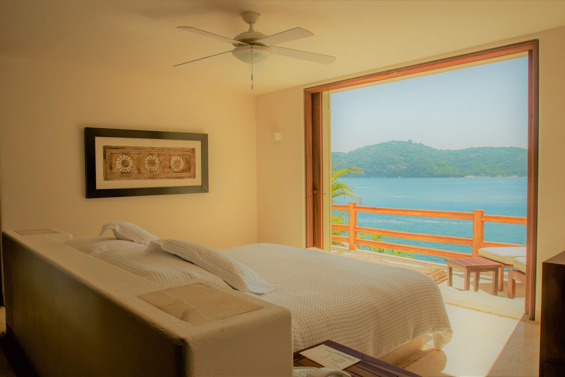 Dormitorio con cama queen, puertas abiertas a un balcón con vista a un océano azul y colinas verdes.