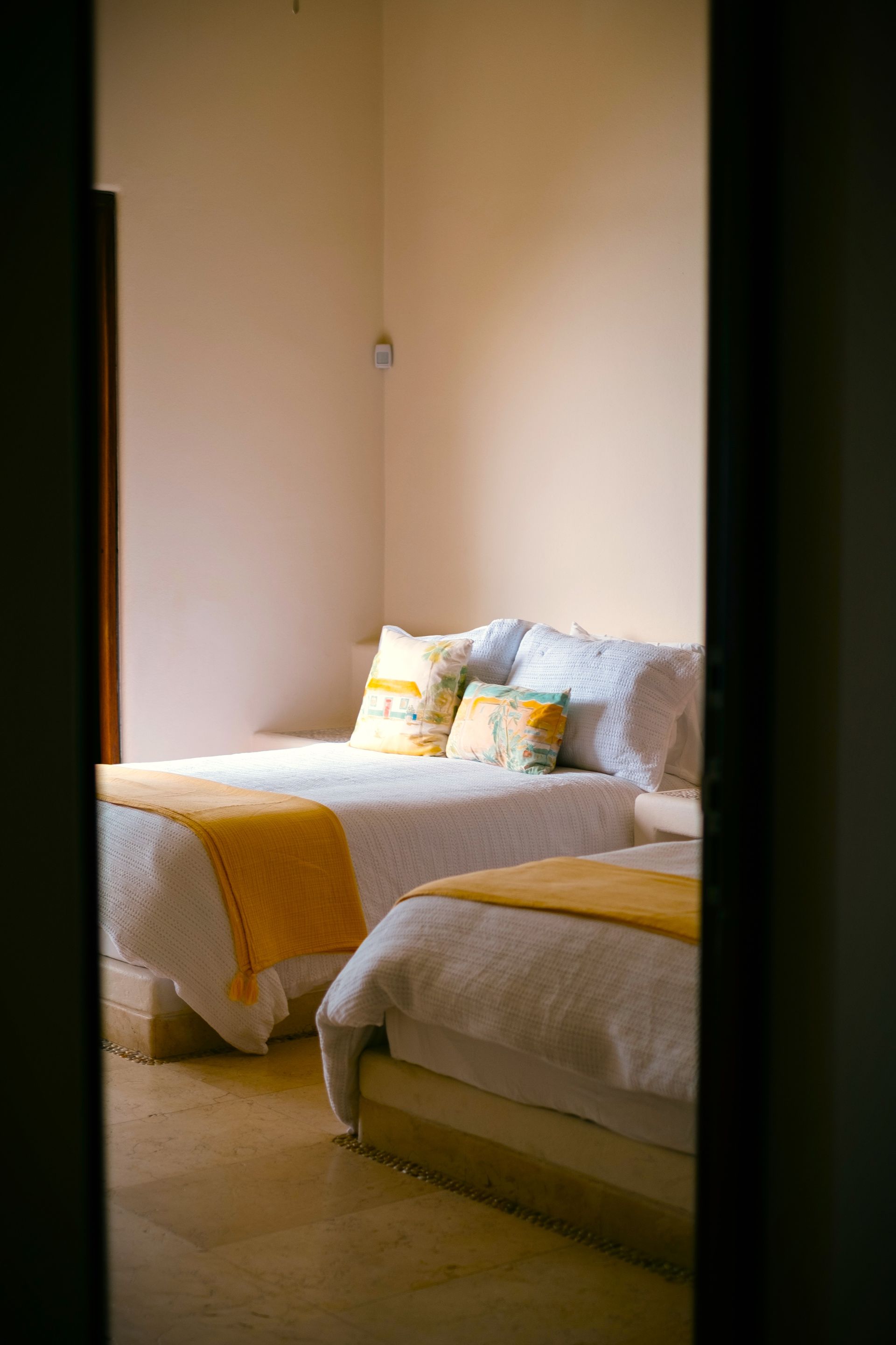 Dos camas individuales en un dormitorio, vistas a través de una puerta. Ropa de cama blanca con detalles en amarillo.