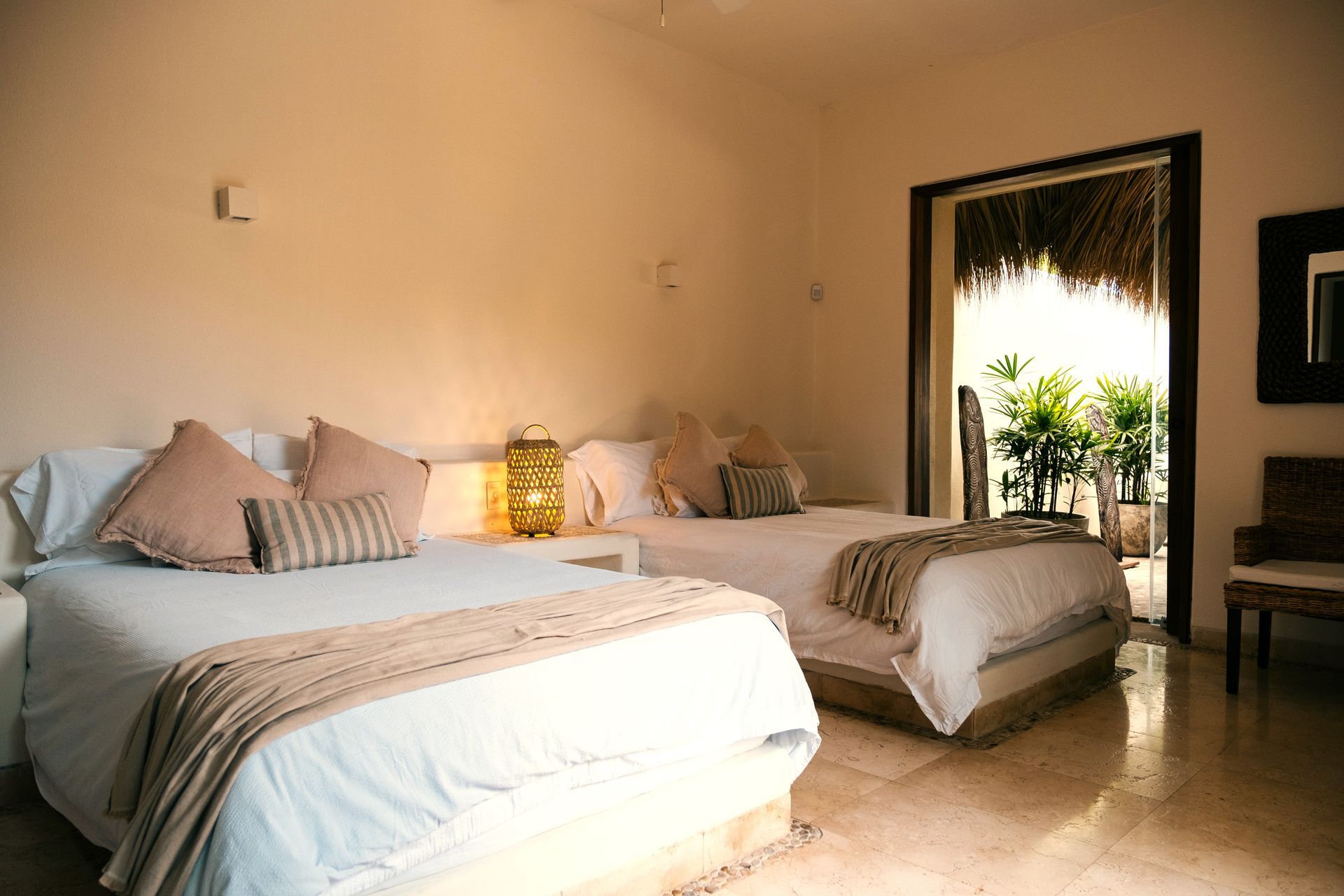 Dormitorio con dos camas y puerta que da a un jardín tropical. Paredes beige y ropa de cama clara.