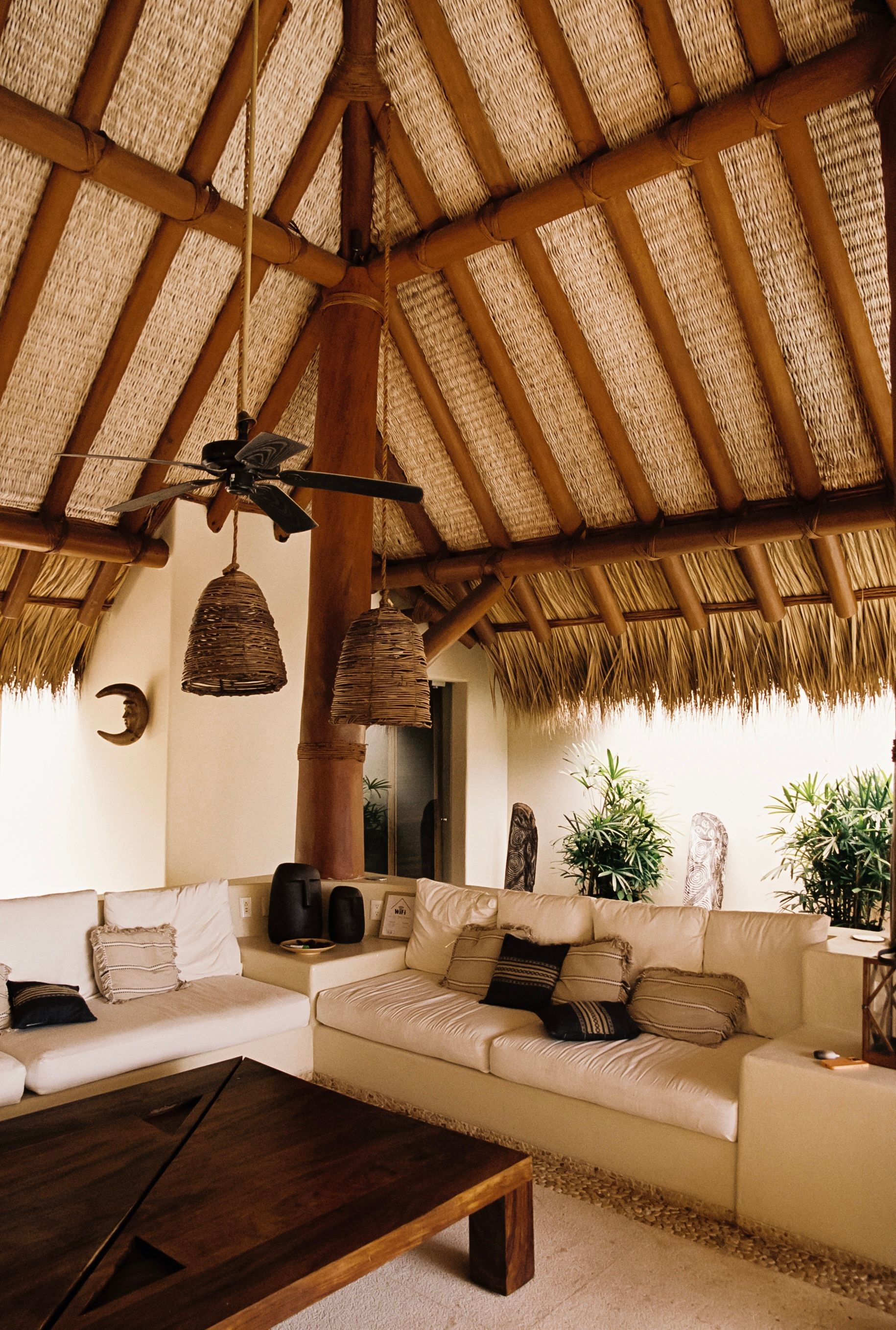 Sala de estar con techo de paja, sofás blancos, mesa de madera y lámparas colgantes.