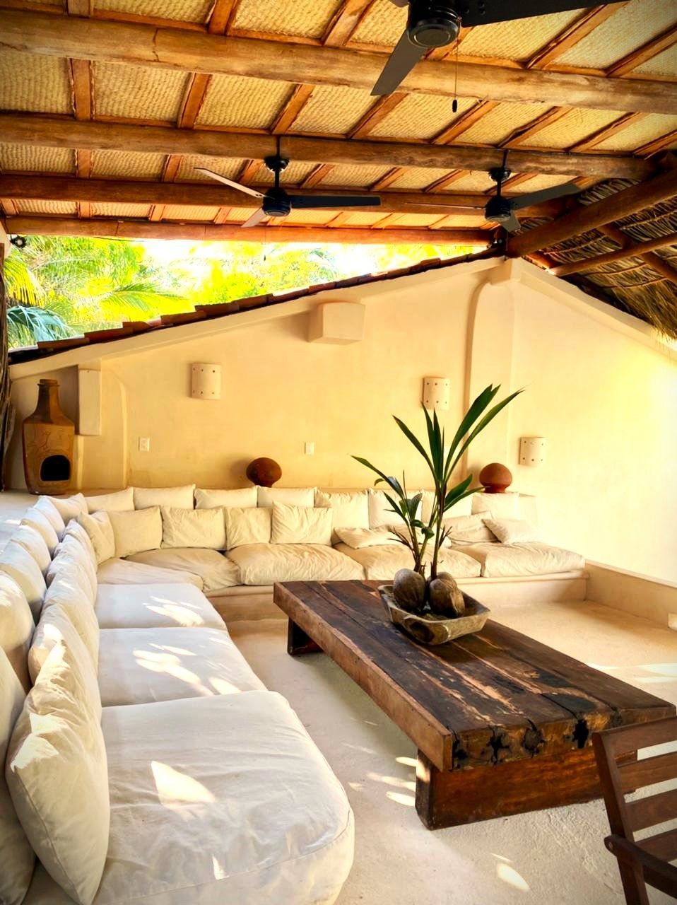 Salón exterior con cojines blancos, mesa de centro de madera, ventiladores y luz natural.