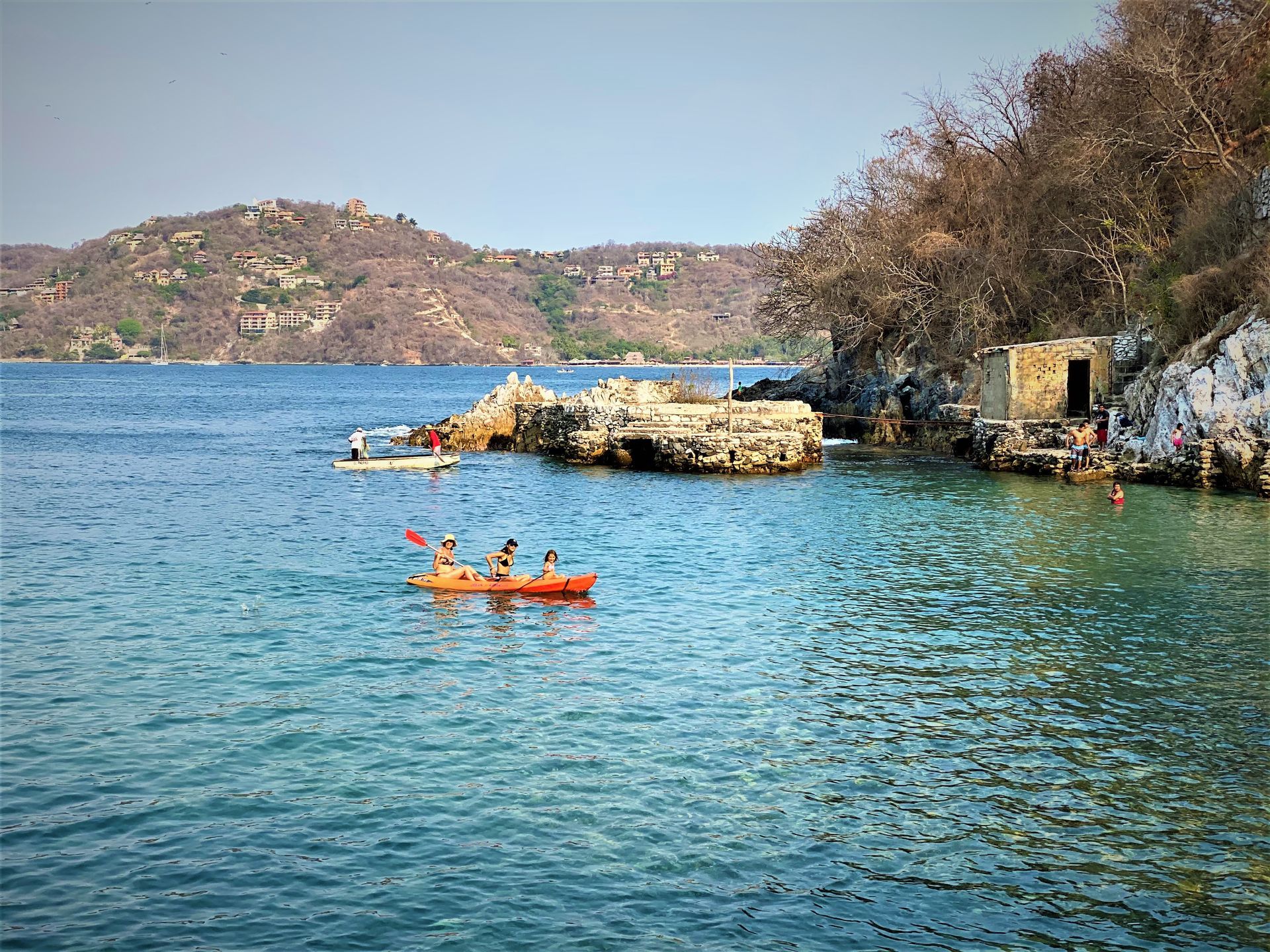 Kayakistas en una bahía azul, costa rocosa y colinas secas bajo un cielo despejado.