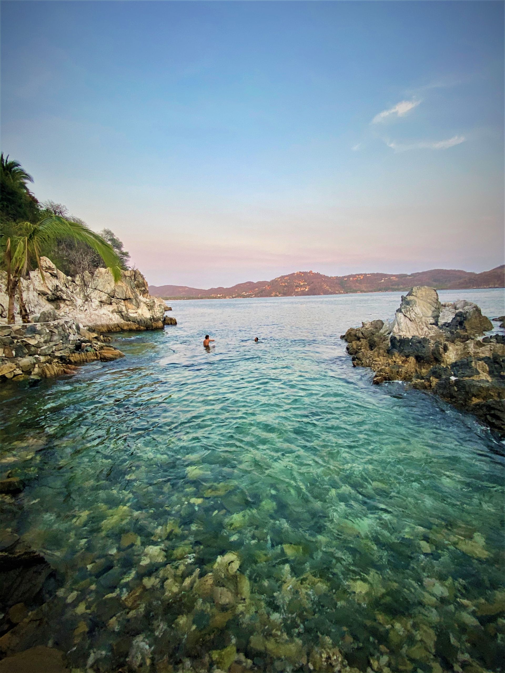 A person swims in a turquoise channel between rocky cliffs under a blue and pink sky.