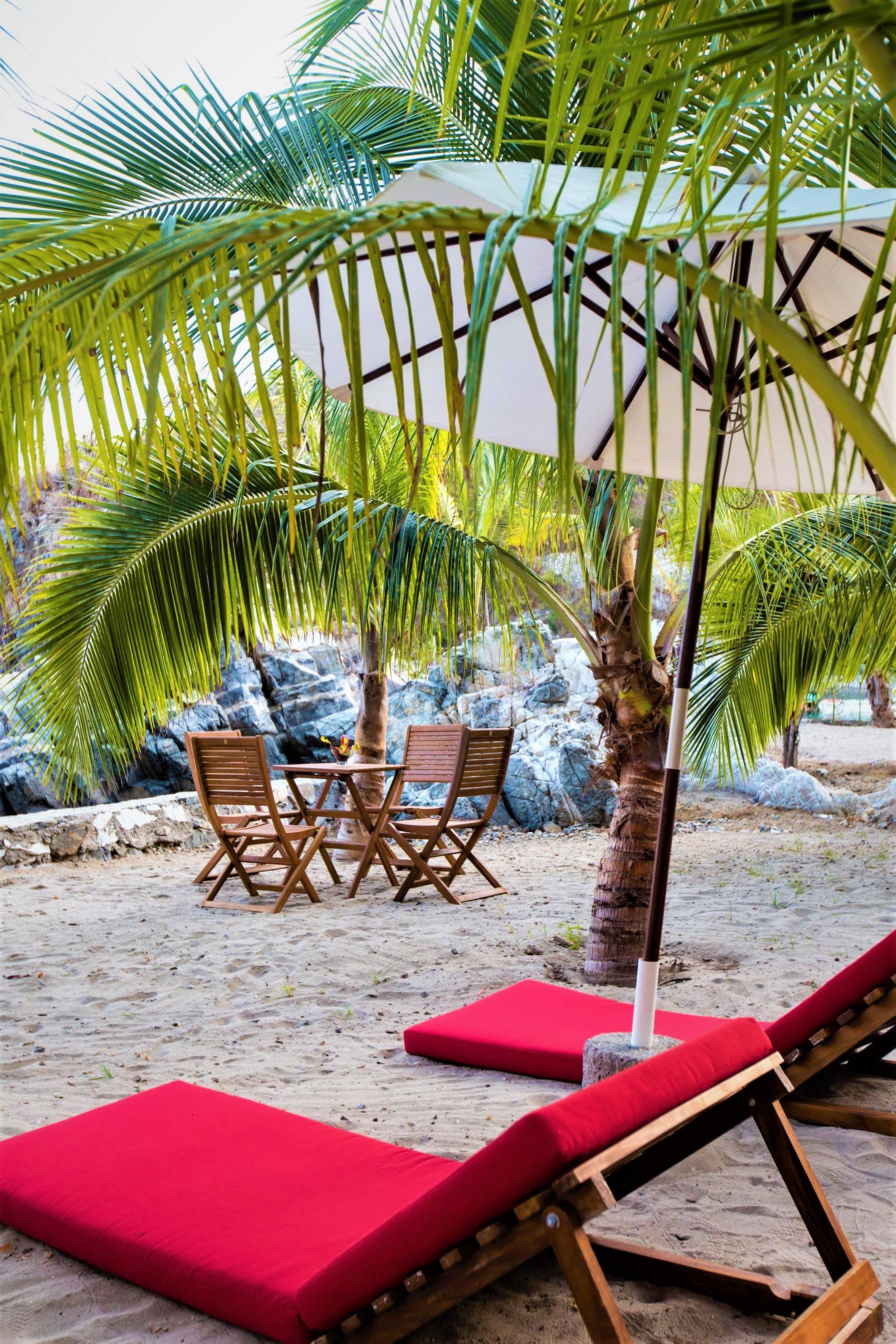 Escena de playa: tumbonas, mesa, sillas, sombrilla bajo palmeras, rojo, arena.