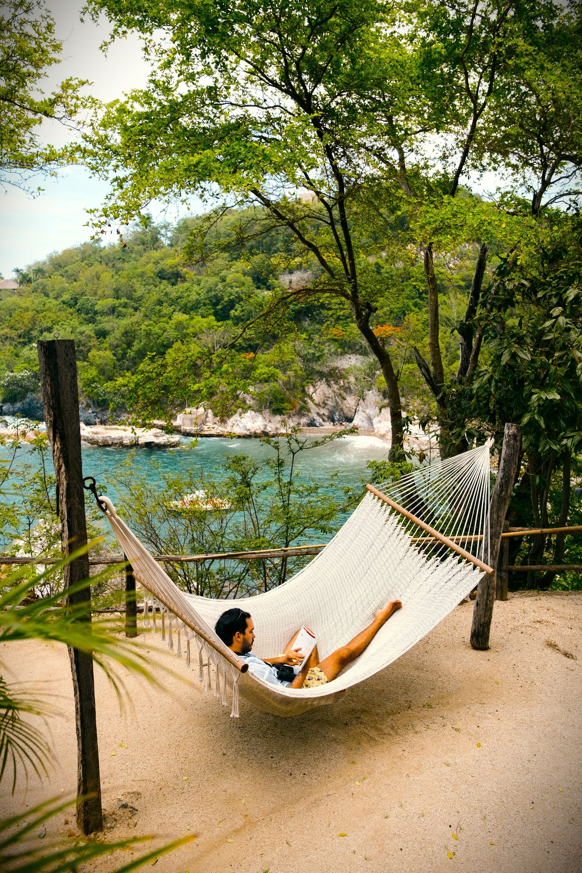 Persona en hamaca, relajándose cerca del océano, con frondosos árboles en el fondo.