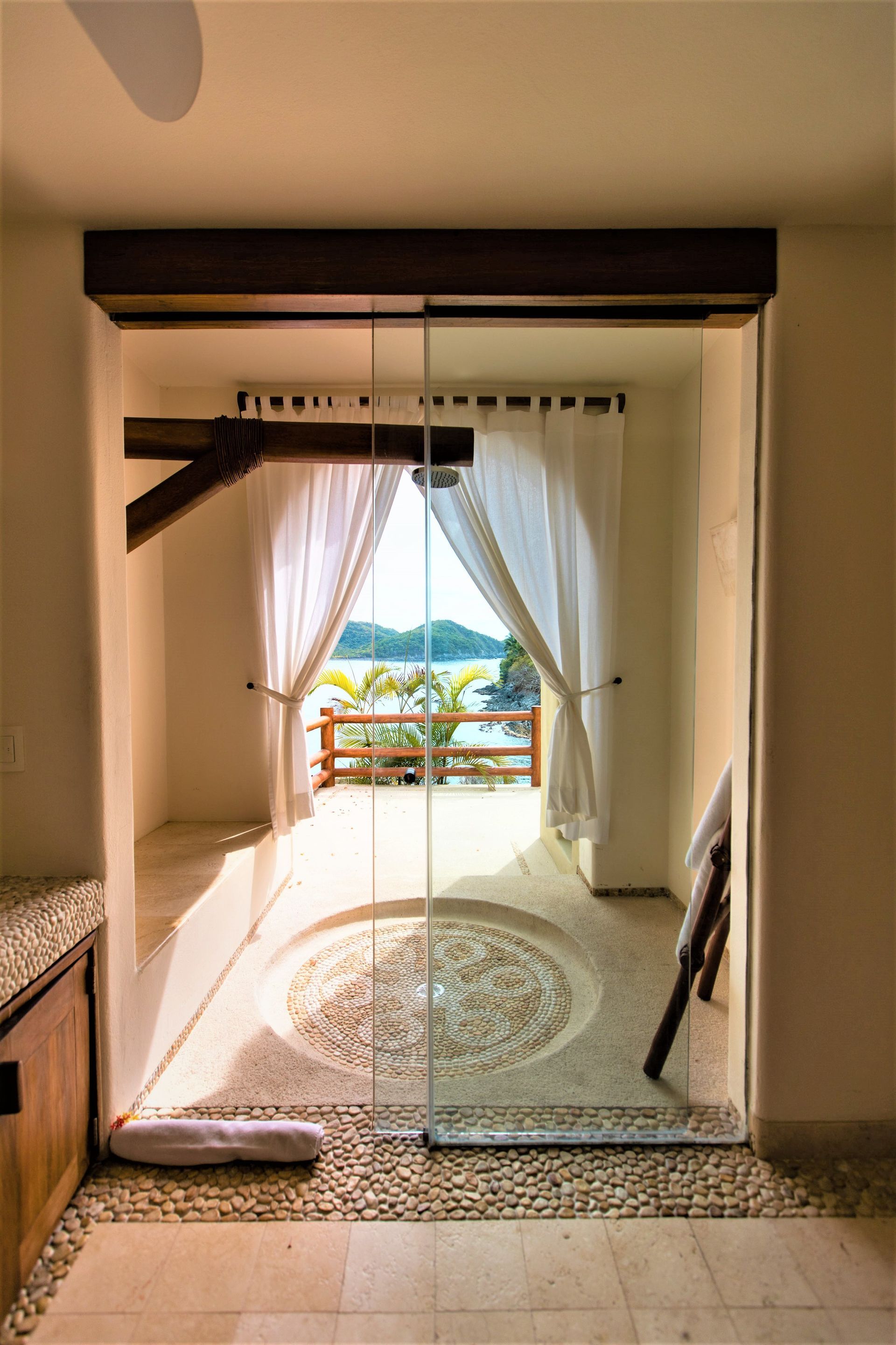 Baño con puerta de vidrio con vista al mar al aire libre, piso y ducha de piedra, cortinas blancas y vigas de madera.