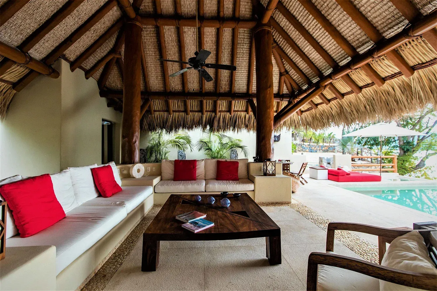Patio cubierto con sofás blancos, cojines rojos y una mesa de café de madera con vista a una piscina.