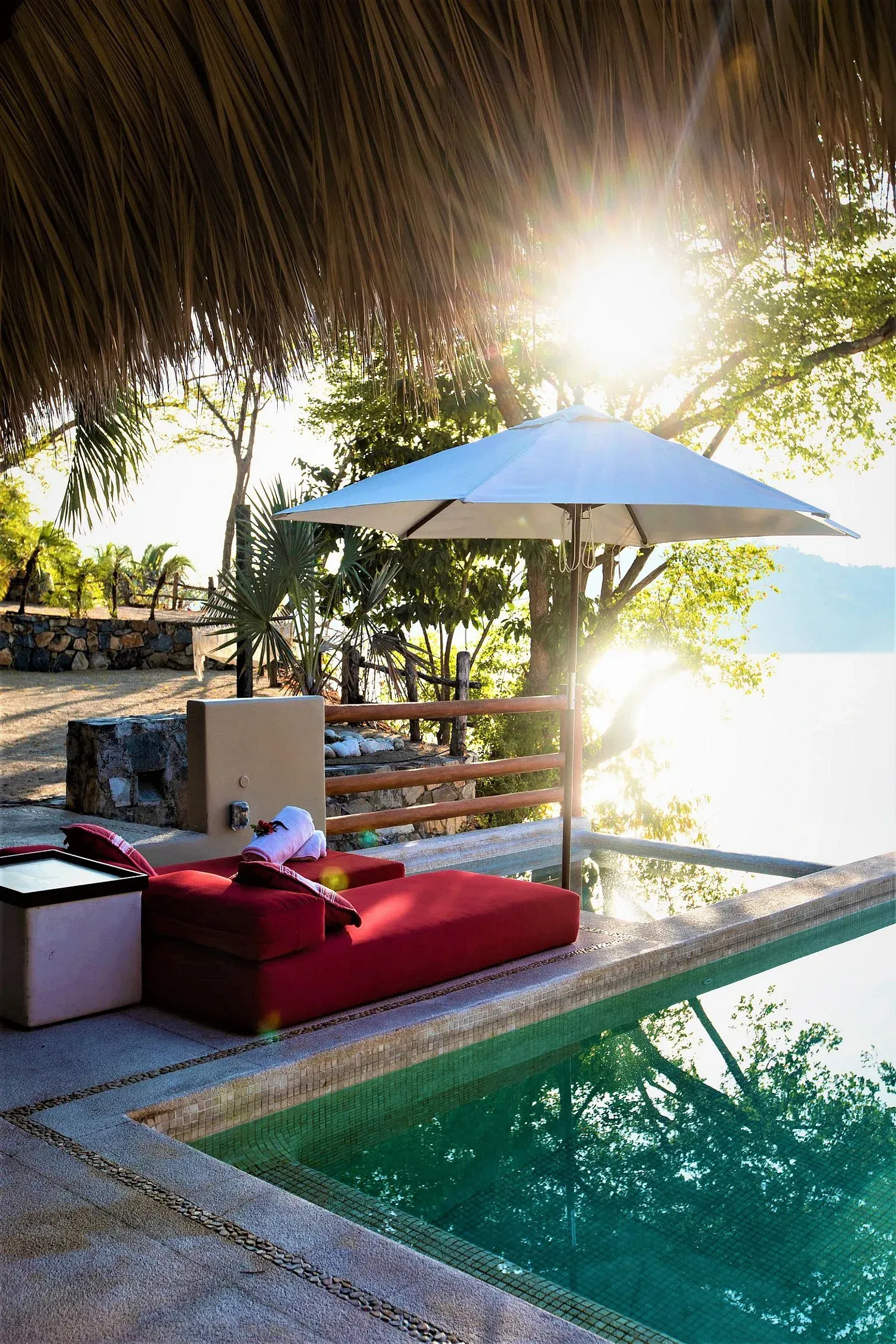 Tumbona roja junto a una piscina con sombrilla, sol brillando a través de los árboles.