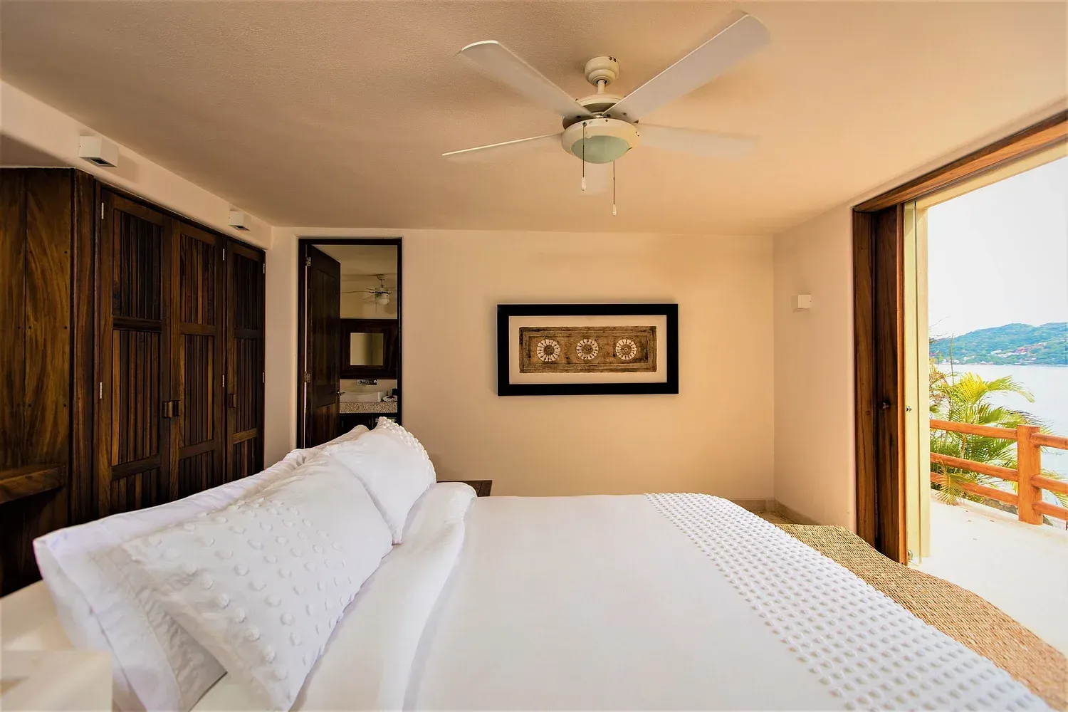 Dormitorio con cama blanca, armario de madera y puertas abiertas a balcón con vista al mar.