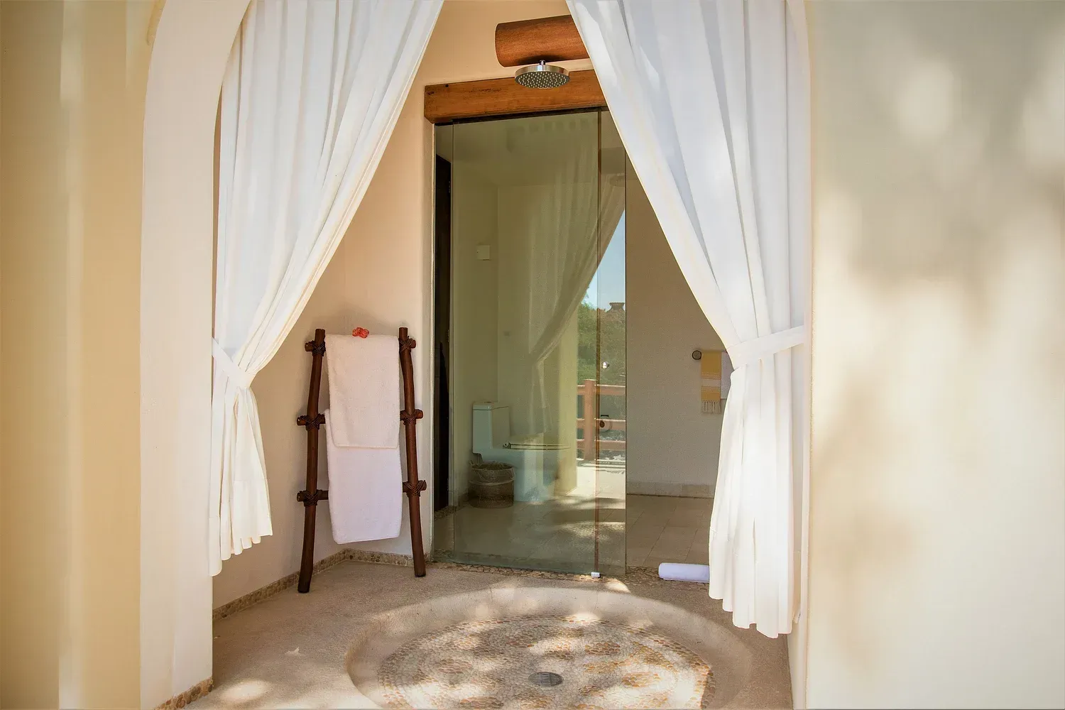 Un baño al aire libre con ducha de cristal, cortinas blancas y escalera para toallas.