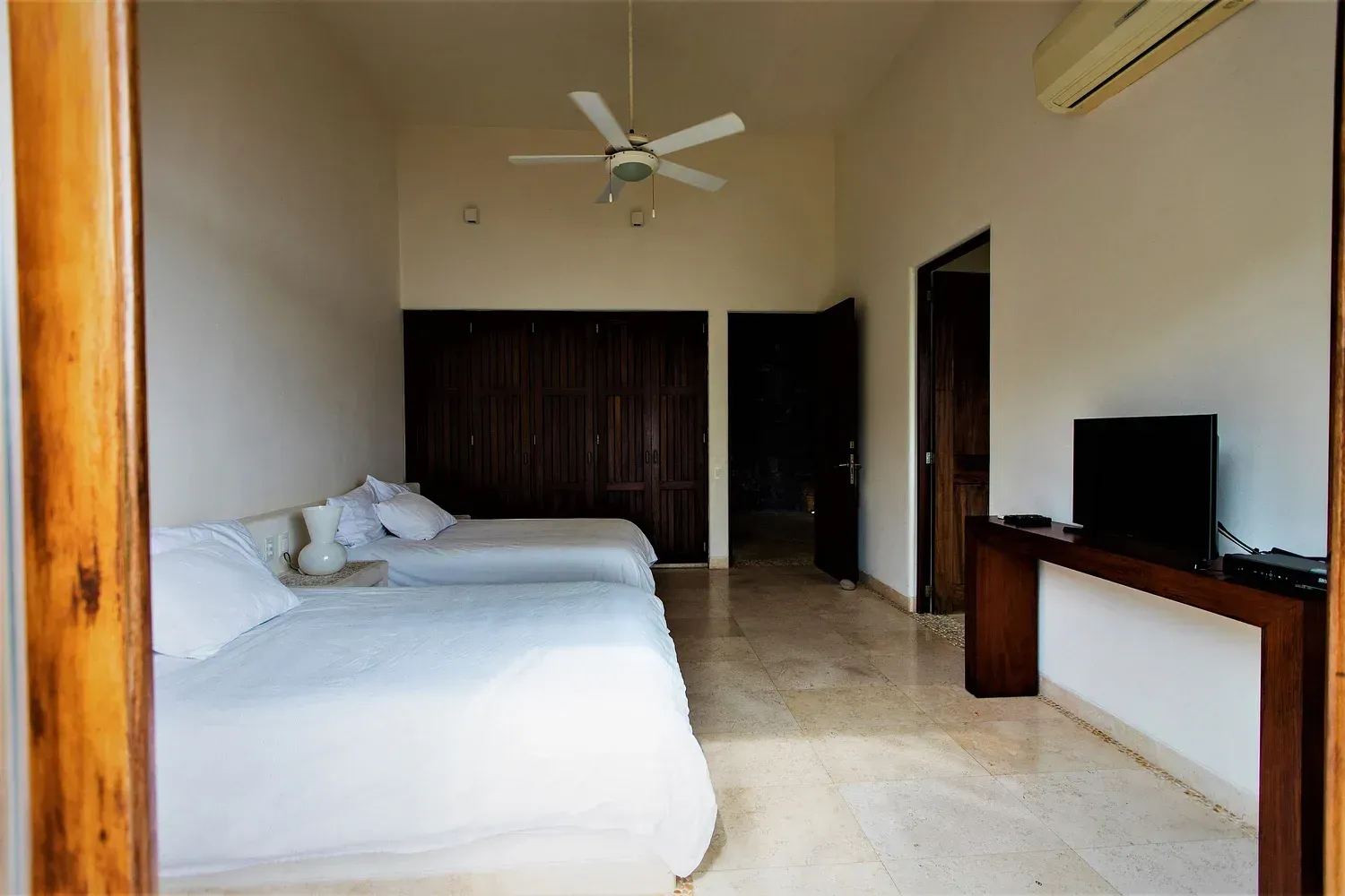 Dormitorio con dos camas individuales, armario de madera oscura, TV y ventilador de techo.