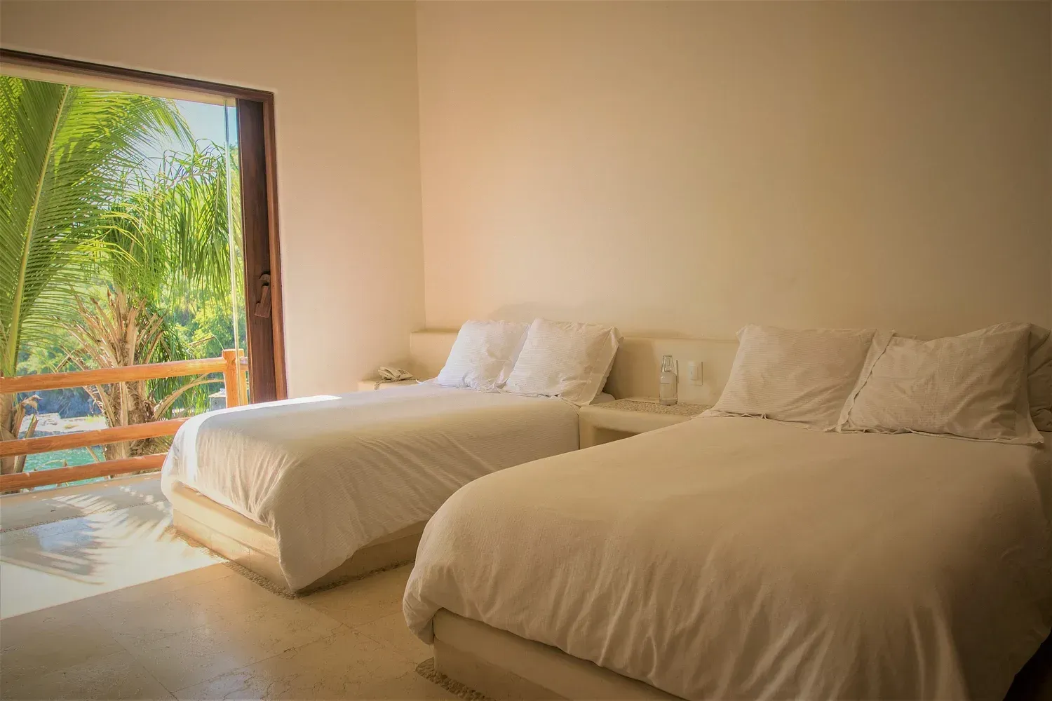 Dos camas individuales con sábanas blancas en una habitación luminosa con una gran puerta abierta a la vegetación.