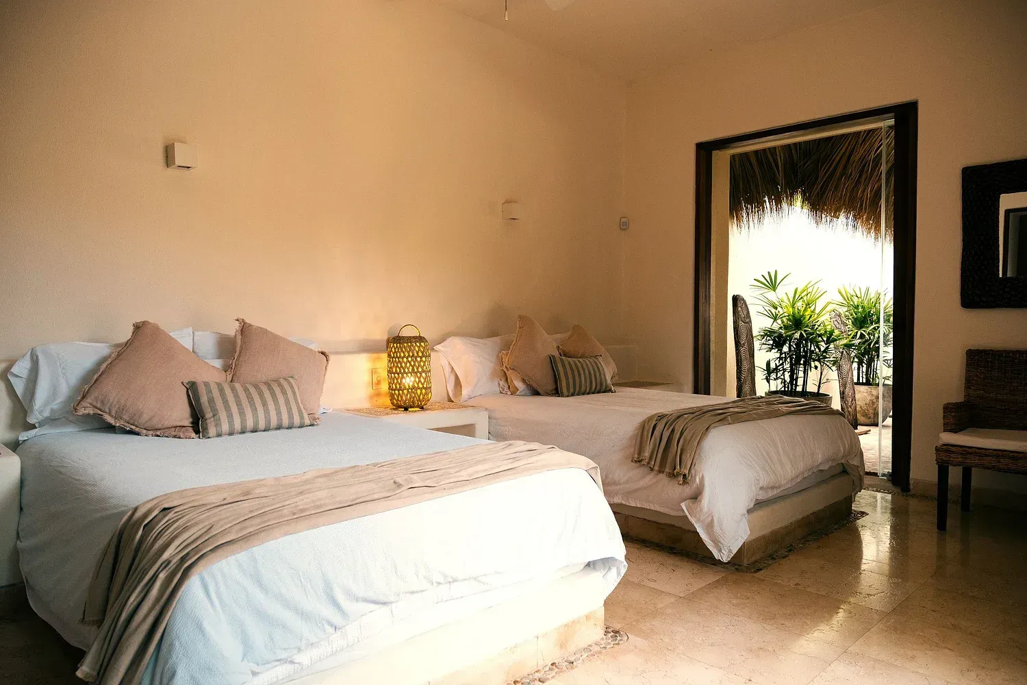 Dormitorio con dos camas, paredes beige y puerta abierta a un patio con plantas.