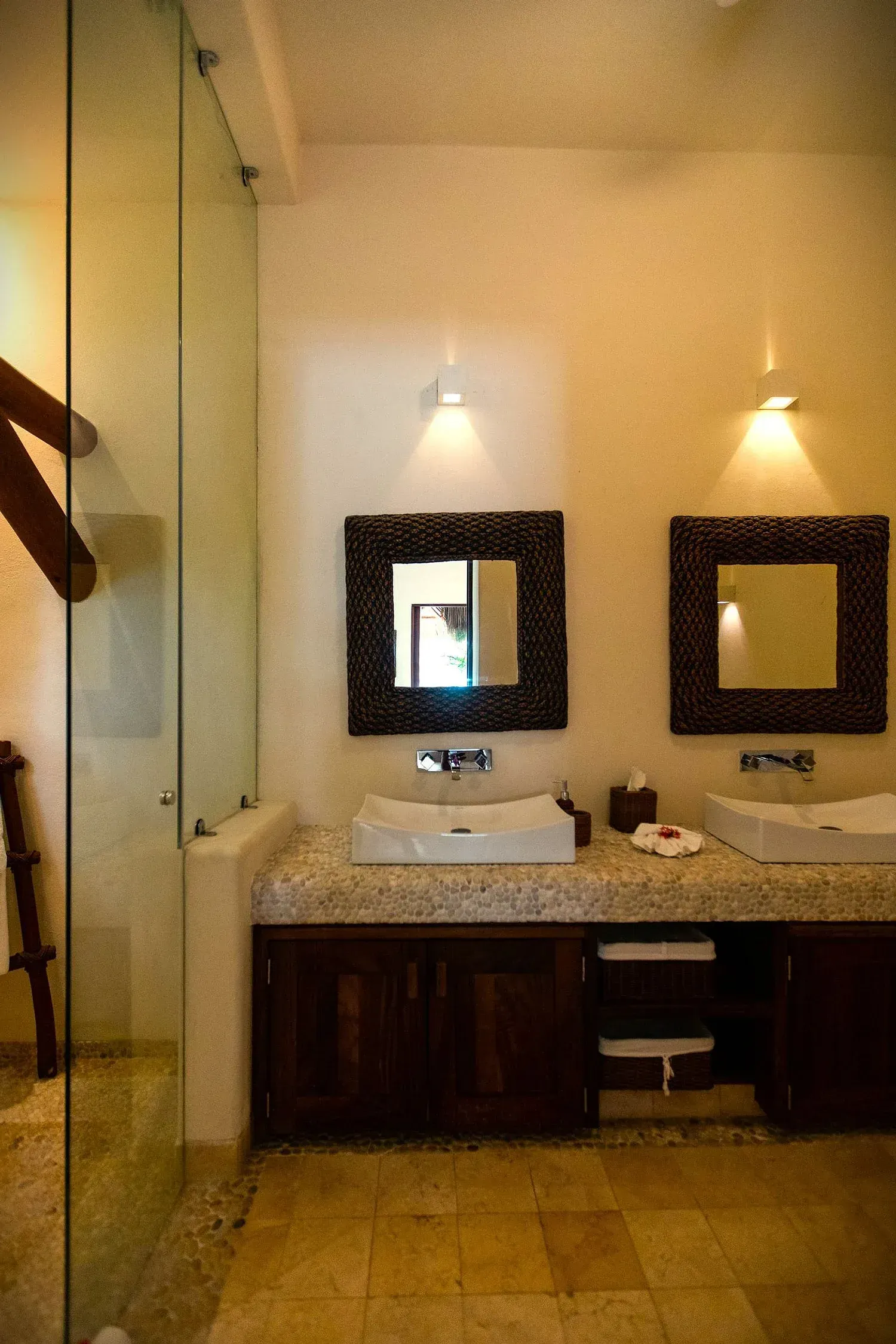 Baño con dos lavabos, muebles de madera oscura, espejos cuadrados y ducha de cristal.