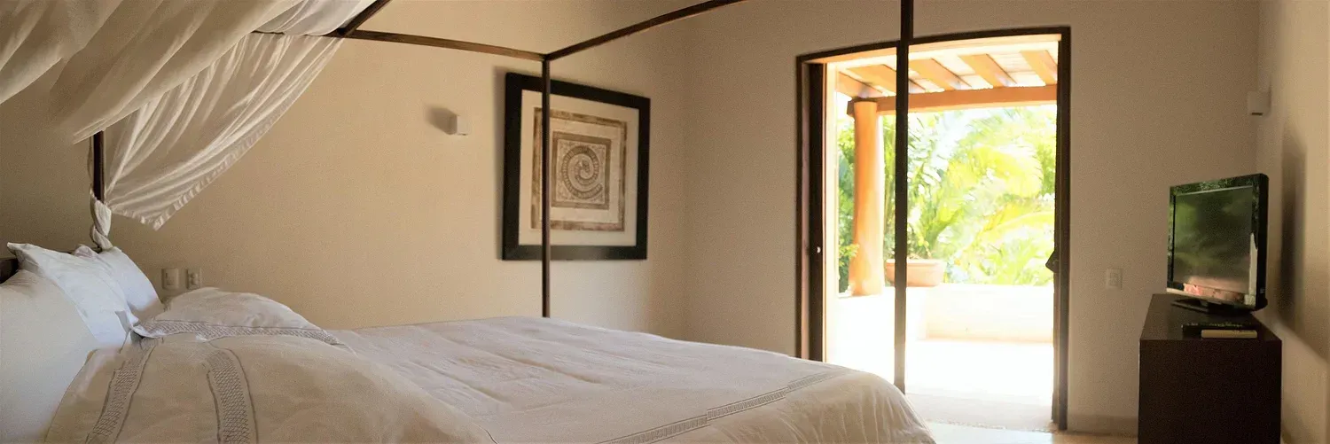 Dormitorio con cama con dosel, obras de arte y puerta corrediza de vidrio al patio con vegetación.