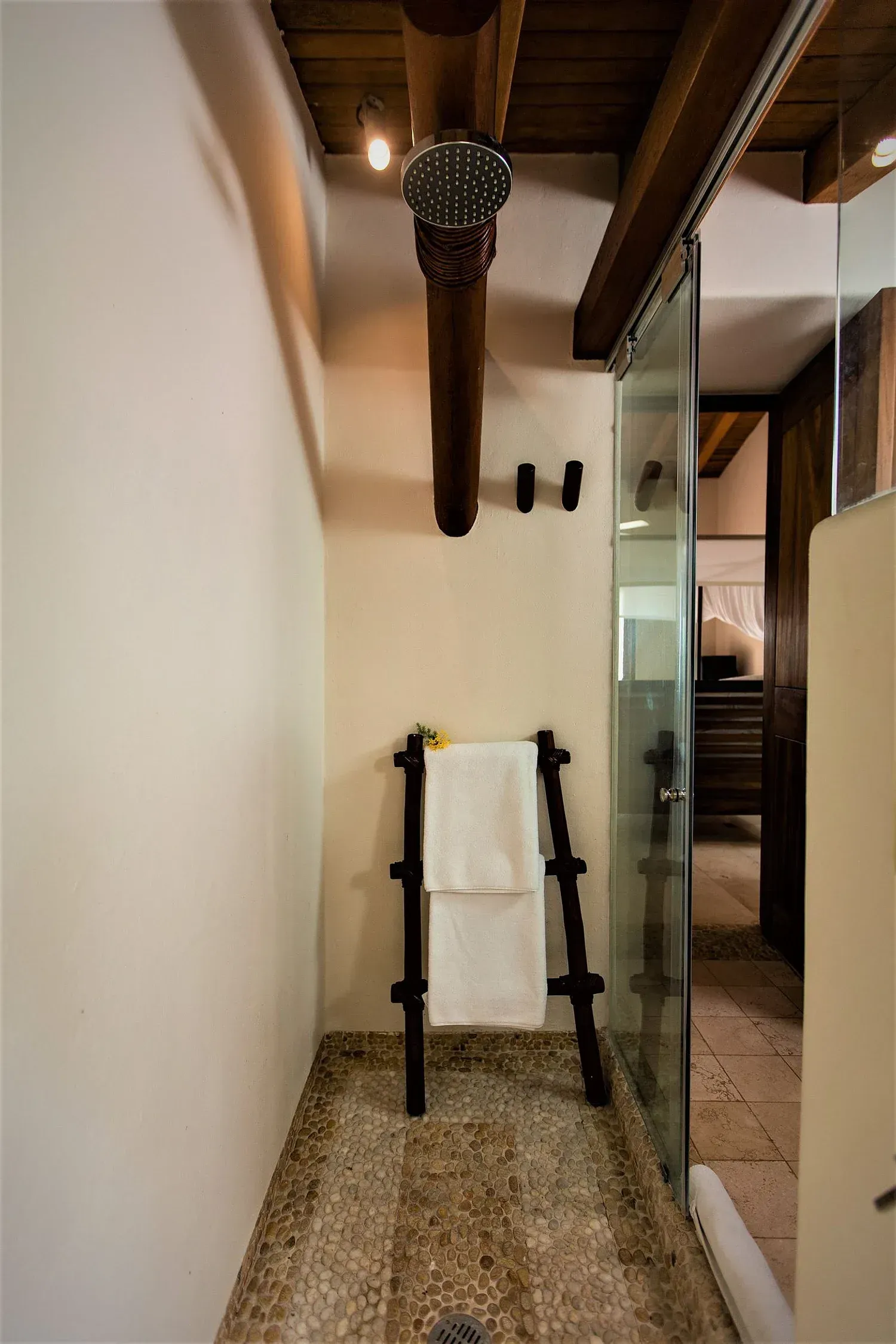 Ducha con suelo de guijarros y escalera para toallas. Techo con vigas de madera y puerta de cristal.