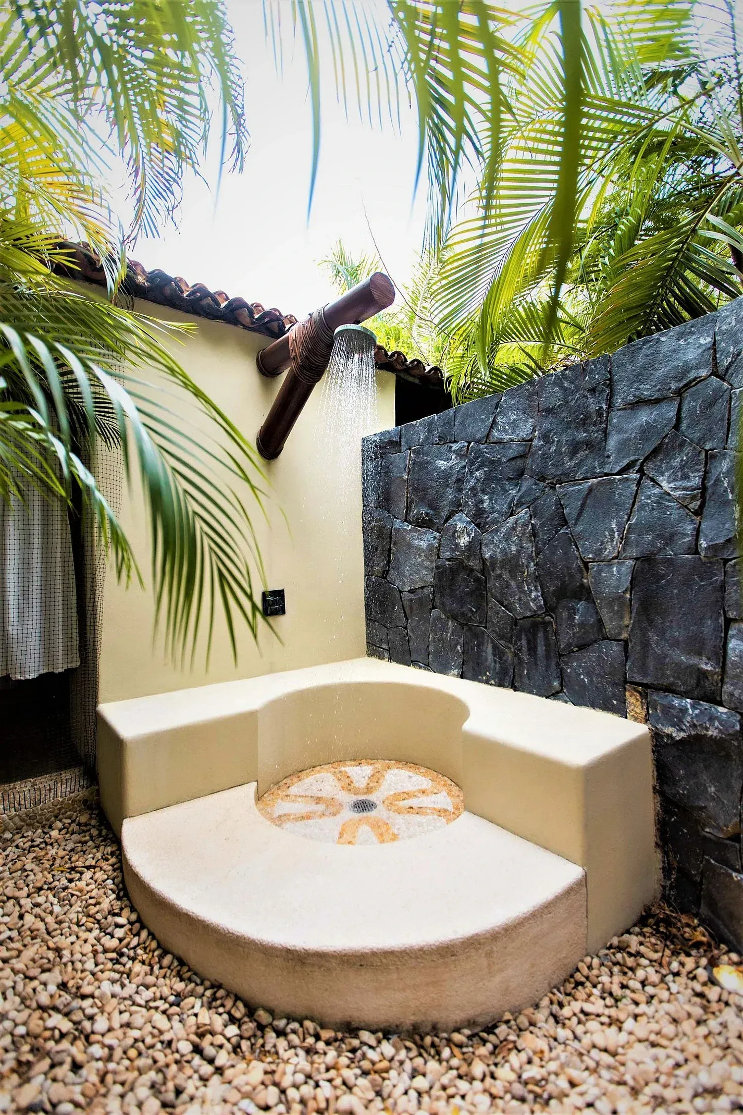 Ducha exterior con piedra y azulejos, rodeada de plantas tropicales.