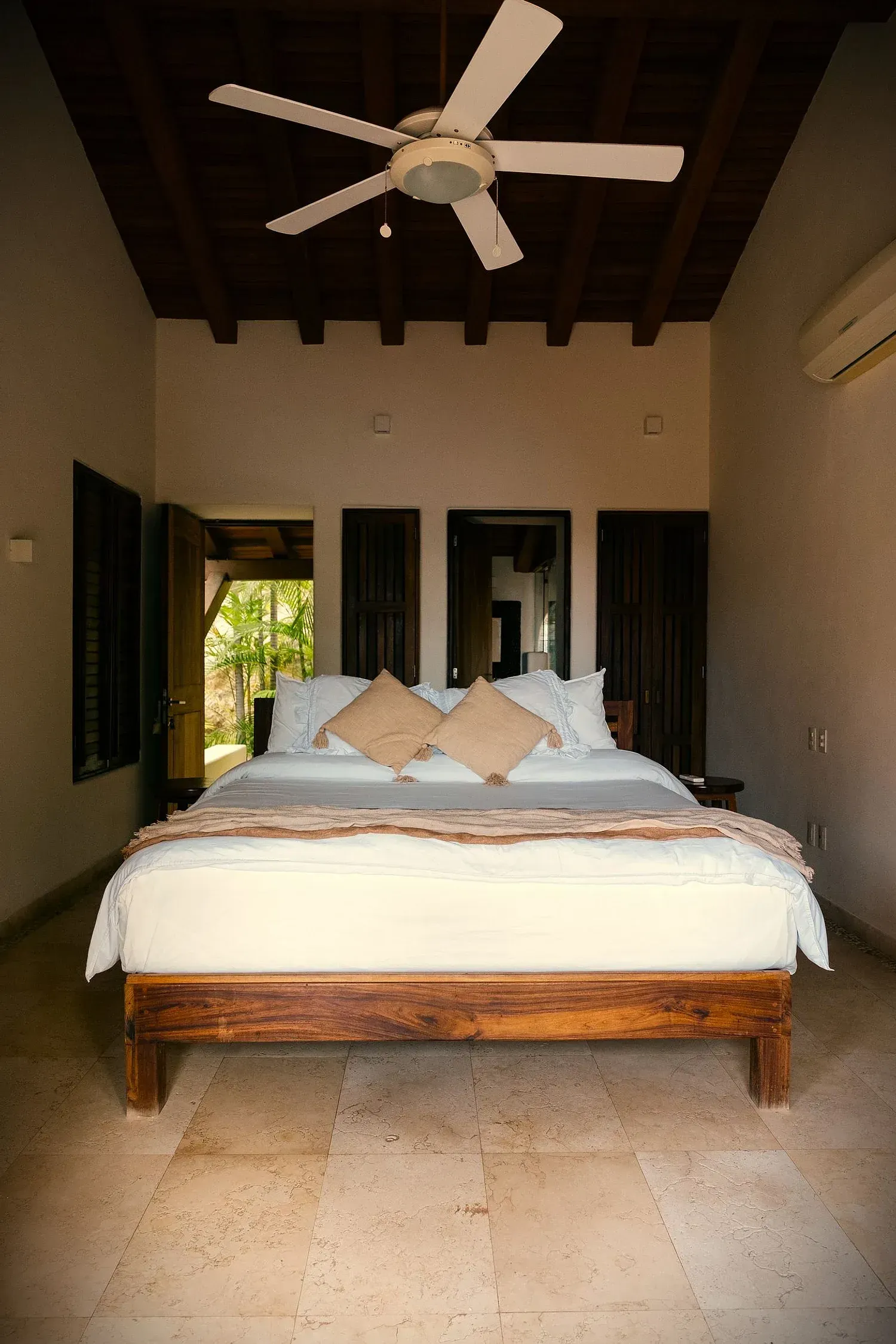 Dormitorio con cama grande de madera, ropa de cama de color claro, ventilador de techo y puertas abiertas.