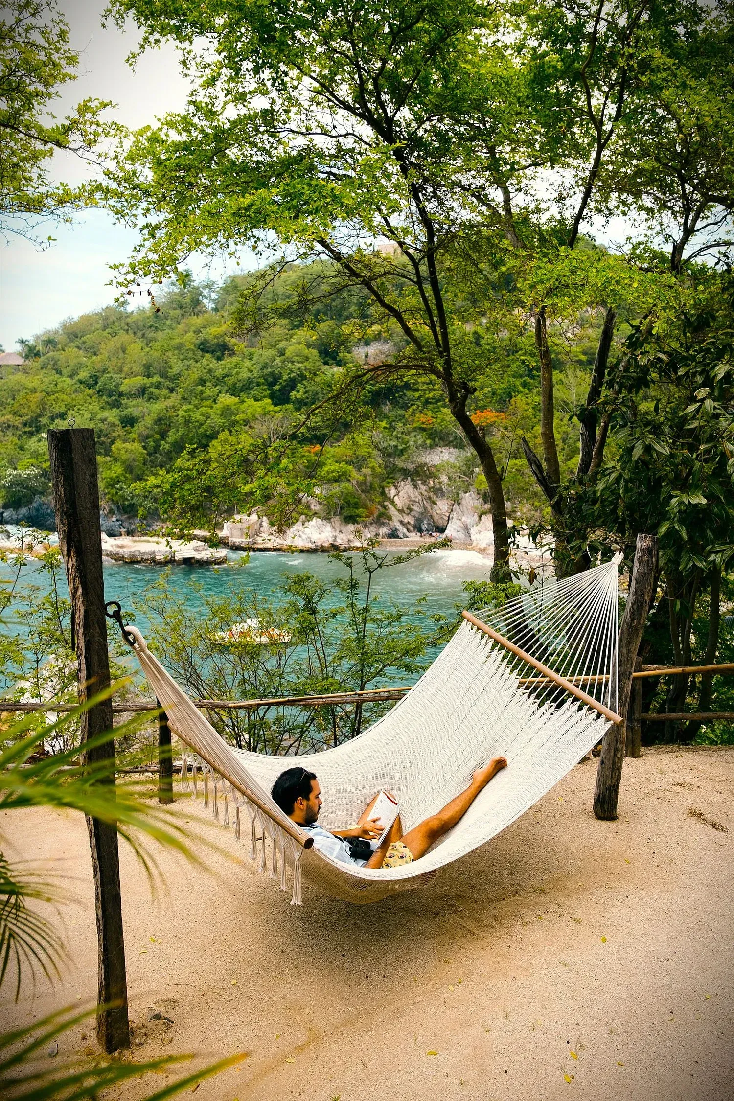 Persona descansando en una hamaca con vista a una bahía, rodeada de árboles.