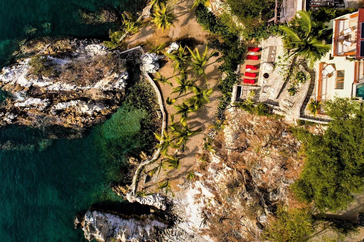 Vista aérea: costa rocosa con agua turquesa, palmeras y un edificio con techo rojo.
