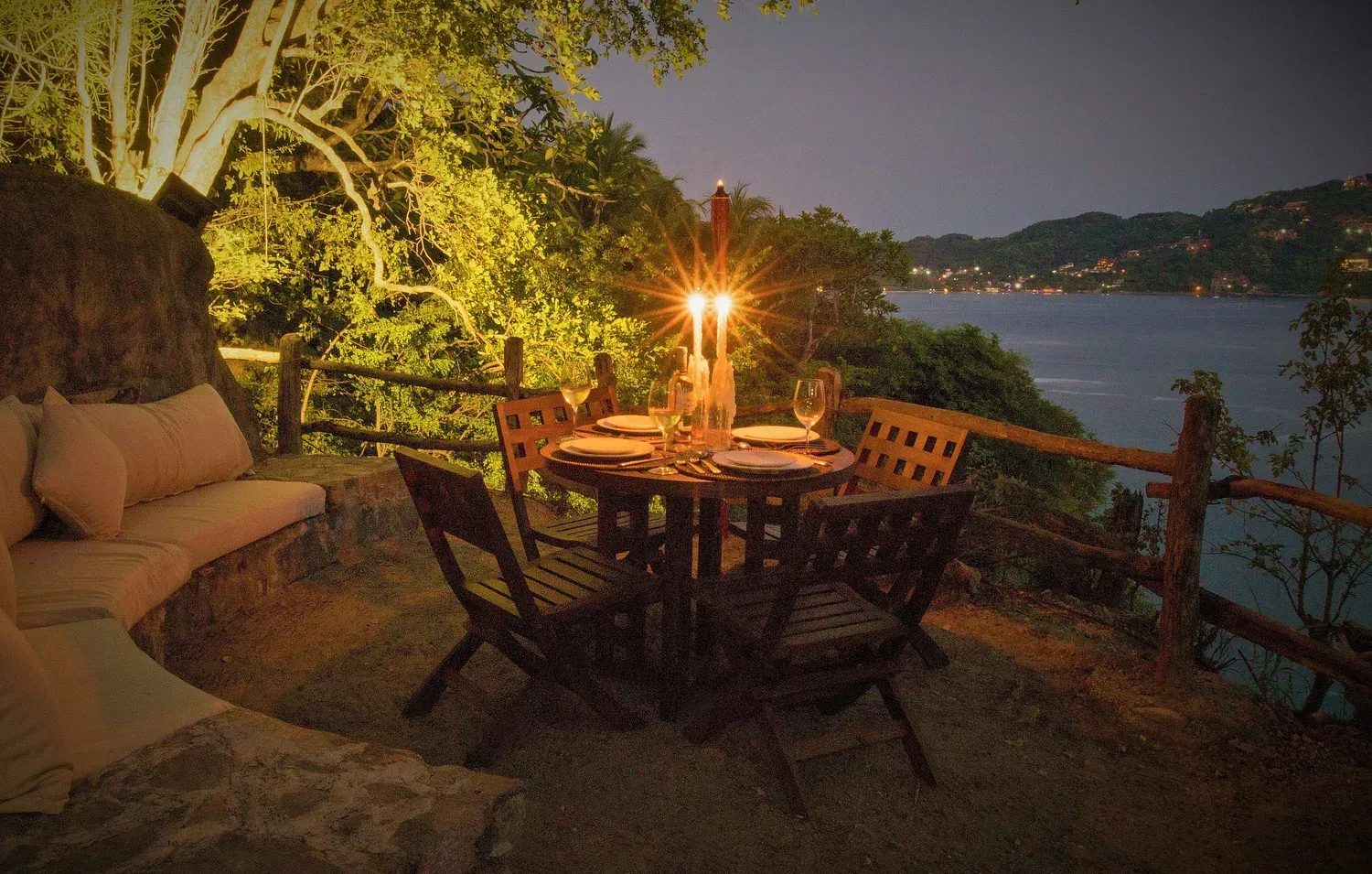 Mesa al aire libre preparada para cenar con velas encendidas, con vistas al agua al anochecer.