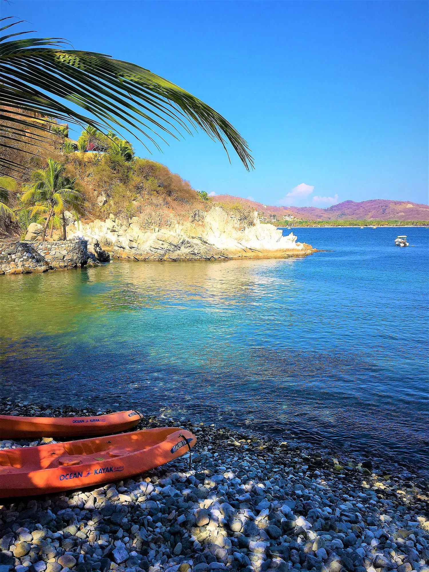 Dos kayaks naranjas en una playa de guijarros junto a aguas cristalinas, costa rocosa, hojas de palmera y cielo azul.